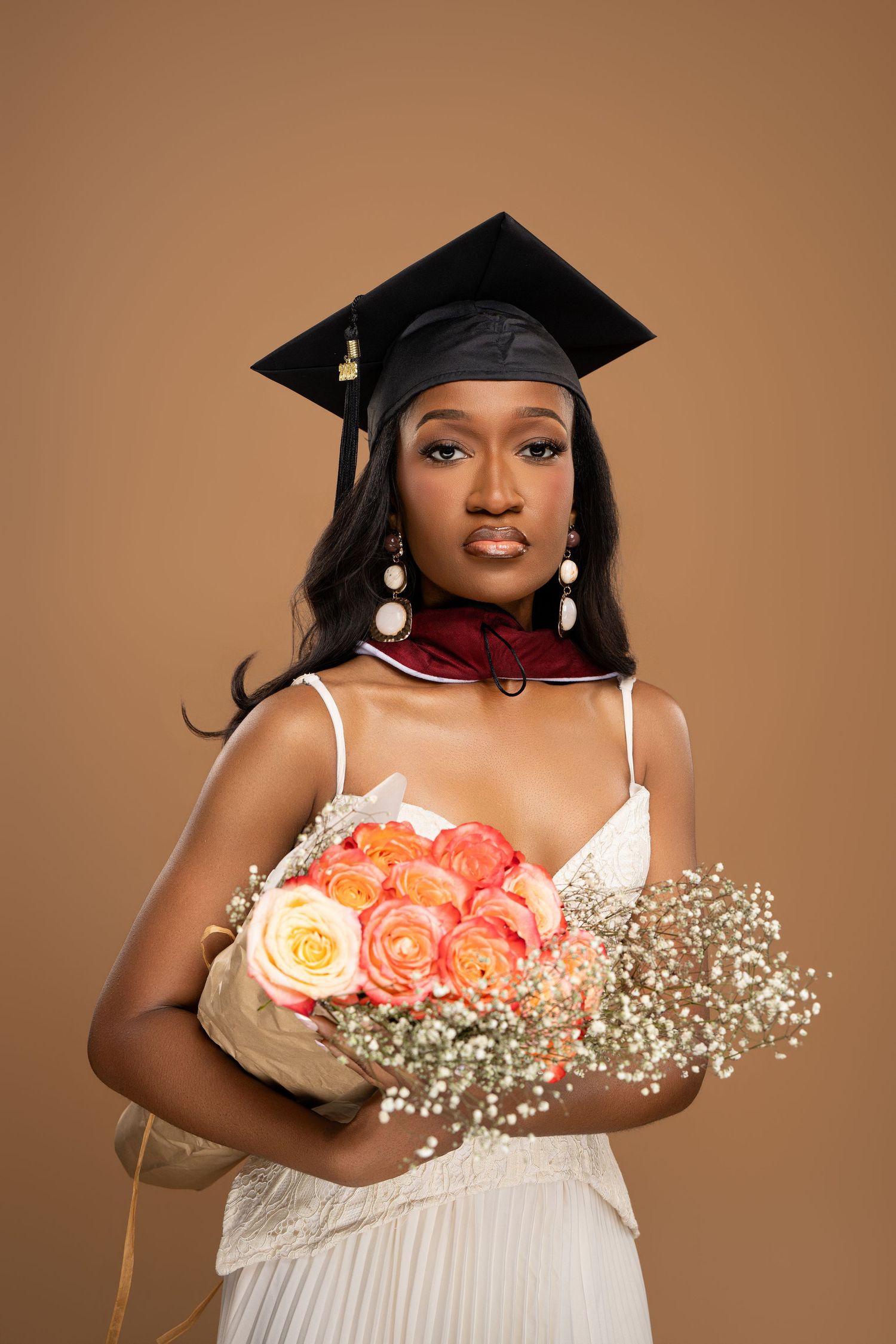 Young Black woman in graduation cap holding peach roses bouquet, wearing white dress and red collar on beige background.