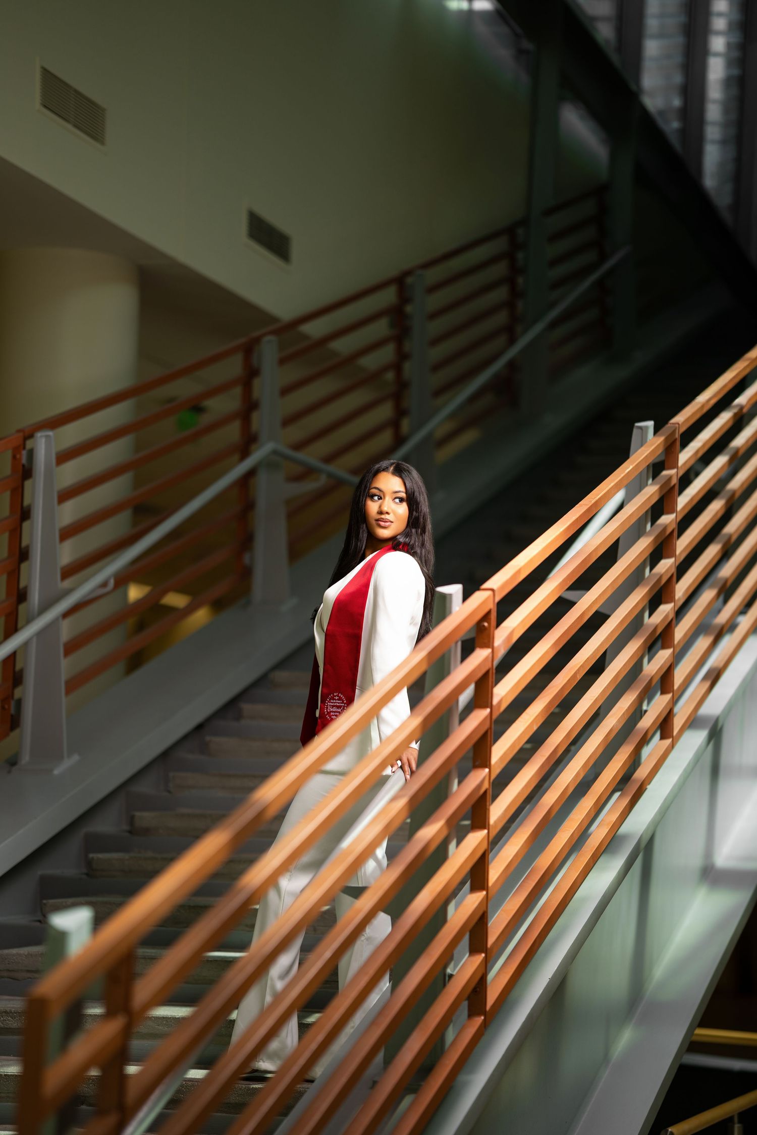 Young woman in red and white graduation stole poses on modern indoor staircase with wooden railings.