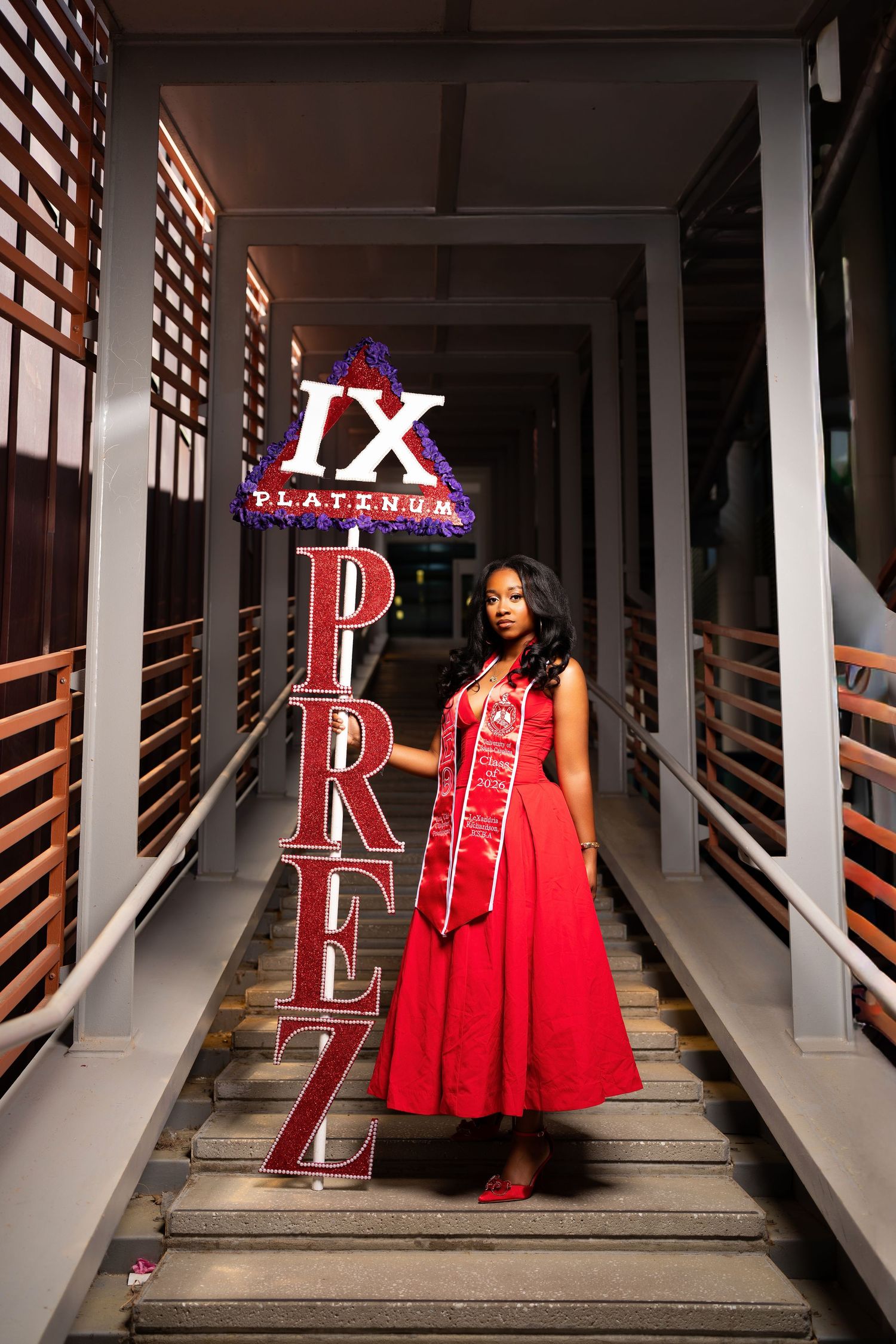 Woman in red gown poses on stairs beside illuminated IX Platinum Prez sign wearing graduation sash.