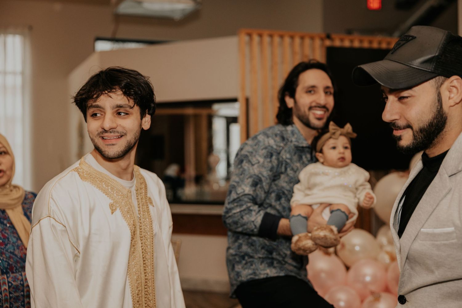 Men smiling at a gathering, one holding a baby in white outfit, another wearing traditional white thobe at indoor event.