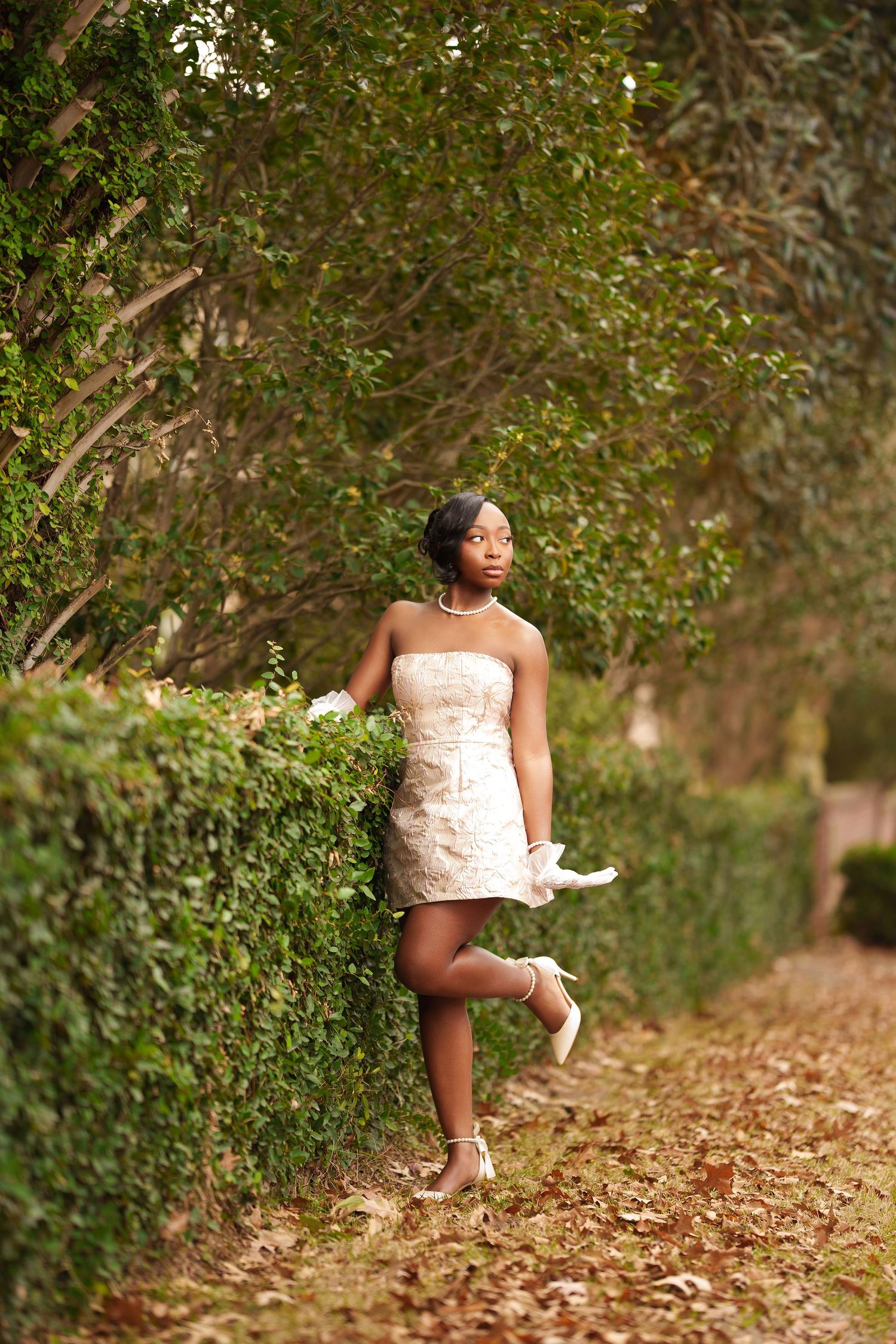 Young Black woman in strapless cream mini dress poses gracefully along a lush green hedge path in an outdoor garden setting.