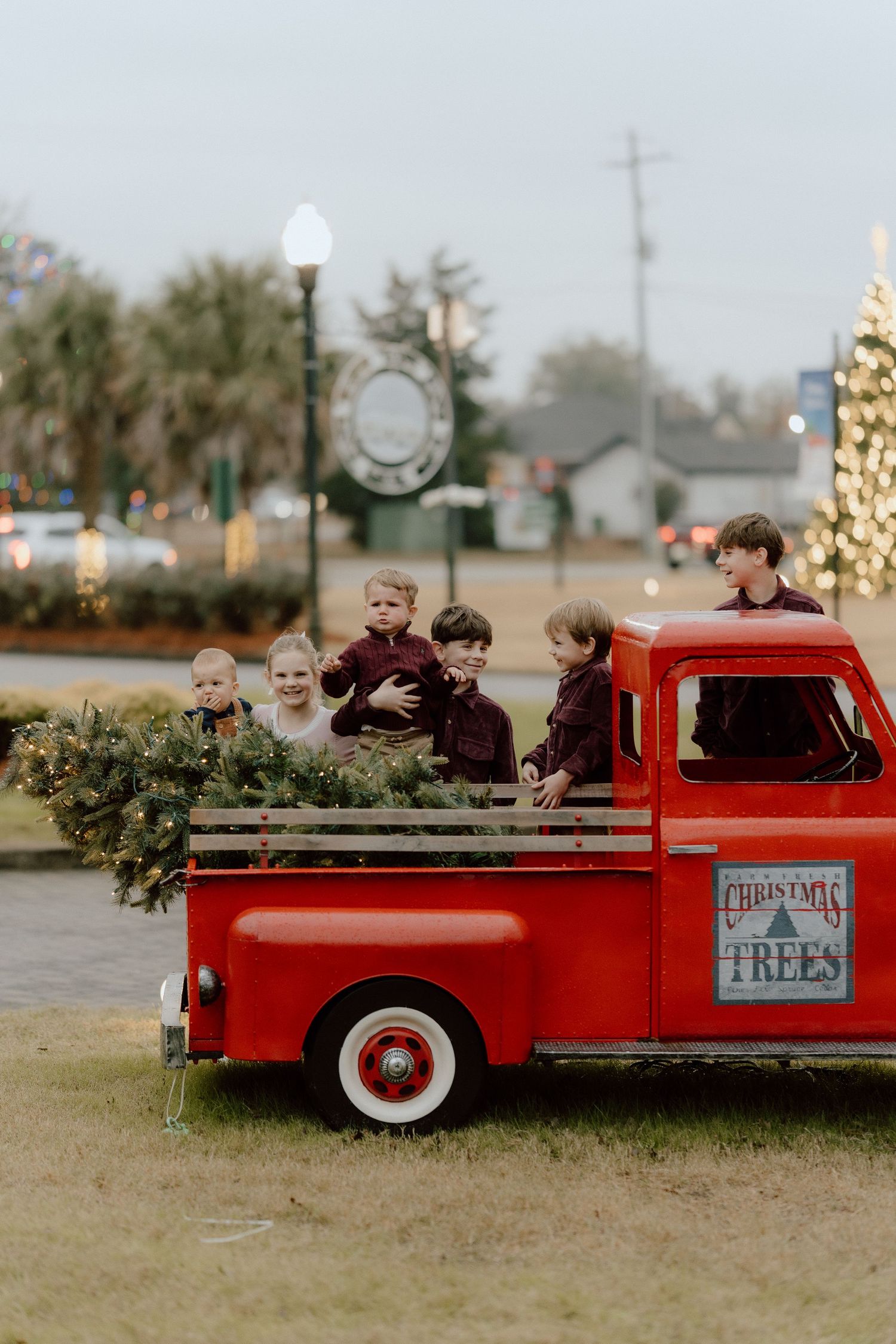 Children ride in the back of a vintage red pickup truck decorated for Christmas tree sales at a festive holiday lot.