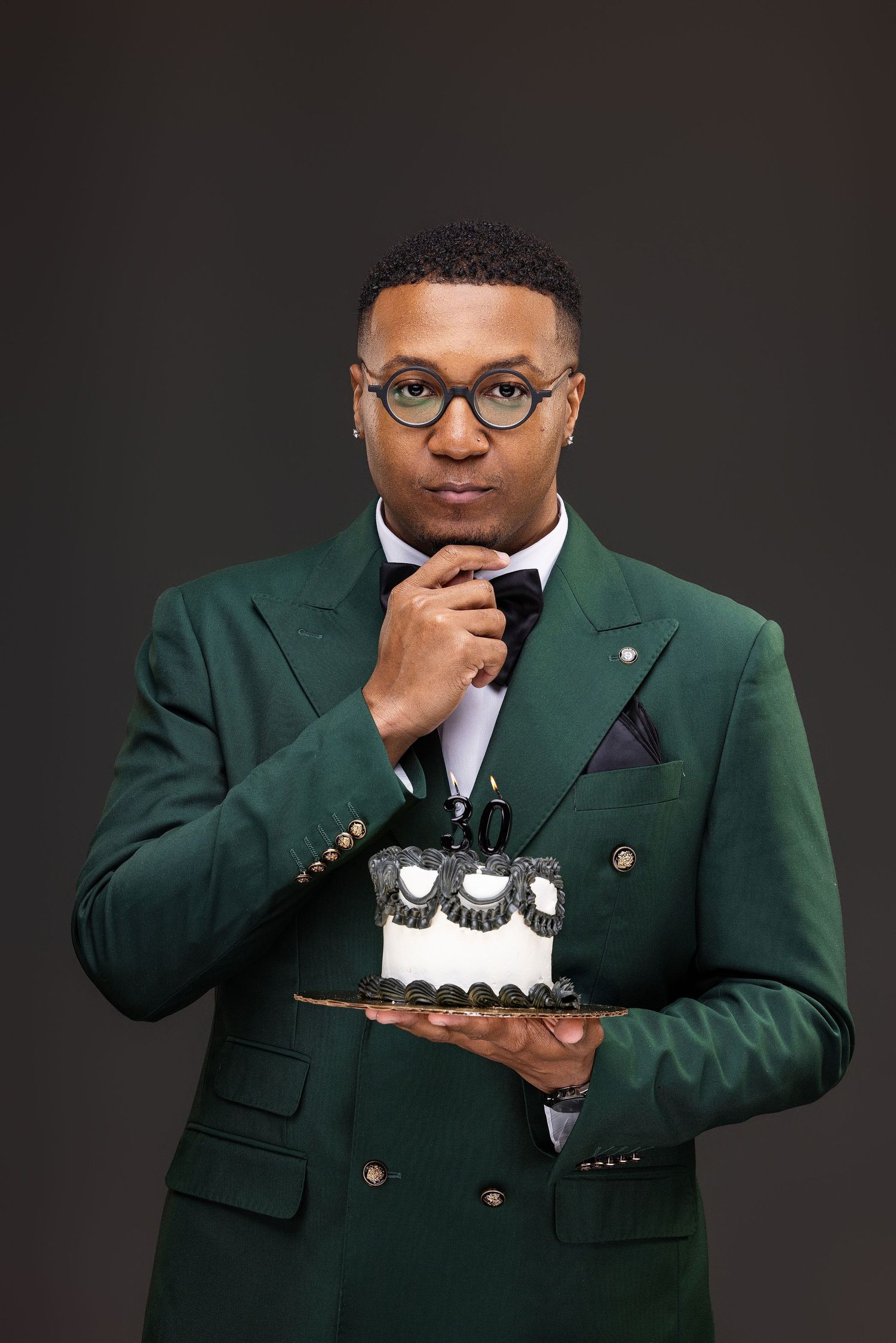 Professional portrait of someone in a green double-breasted blazer and glasses holding decorative cupcakes on a plate.