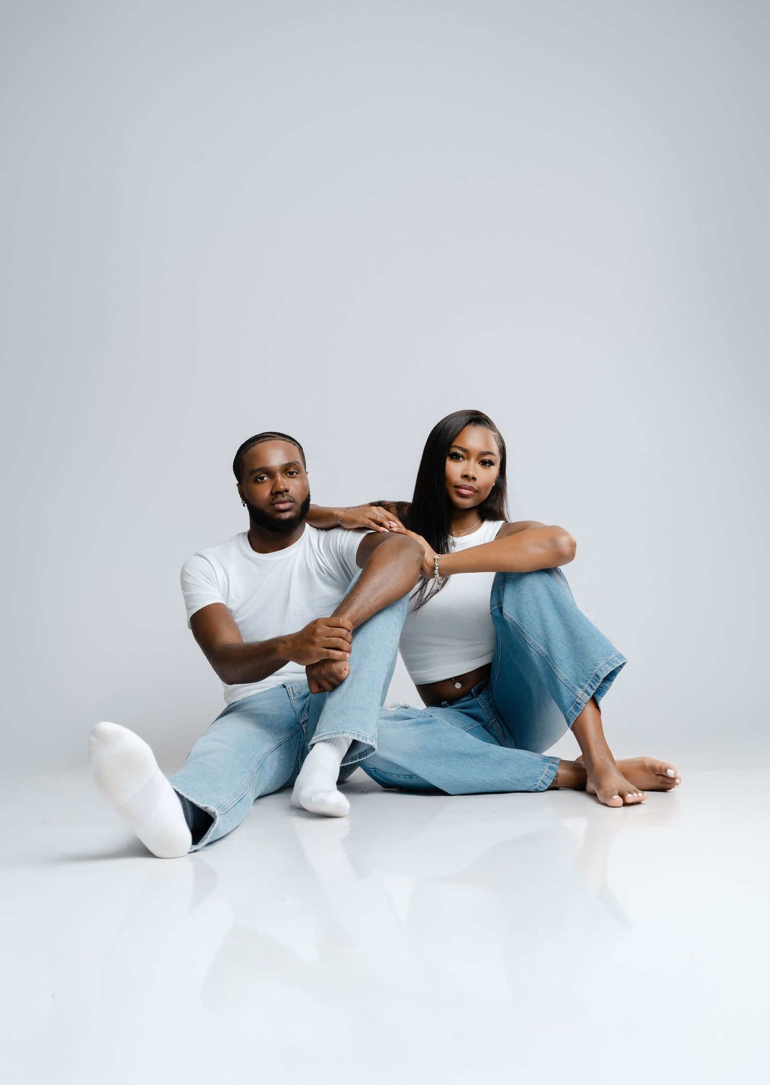 Two young Black adults in white tops and jeans sit together on a white studio floor in a stylish couple portrait.