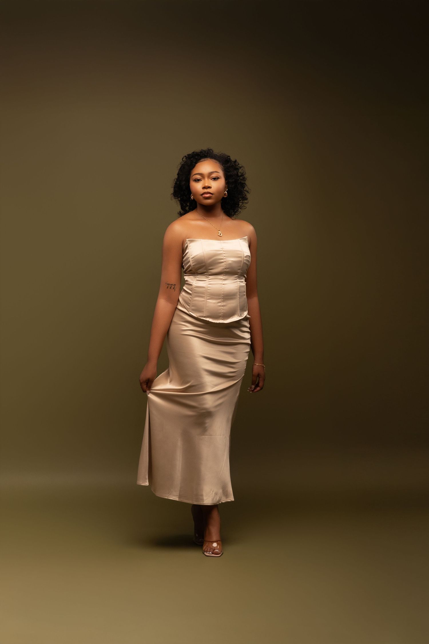 Full length fashion portrait in champagne satin dress against dark brown background.
