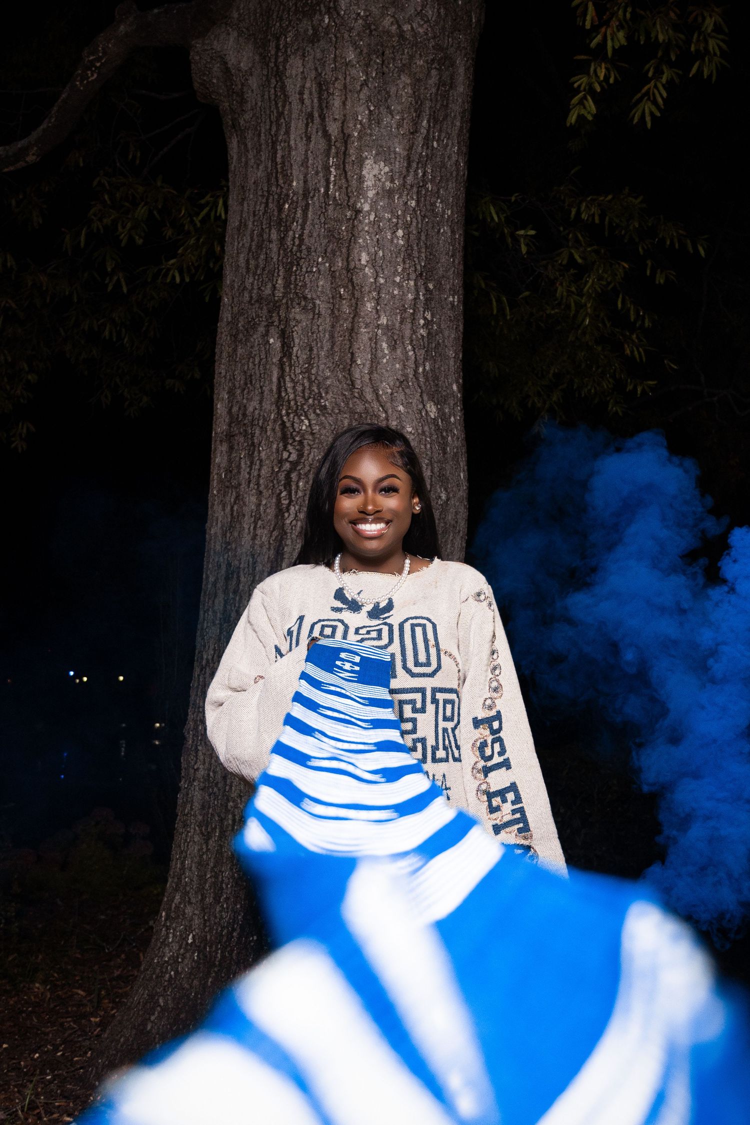 Person wearing collegiate sweater poses by tree trunk with blue smoke effects.