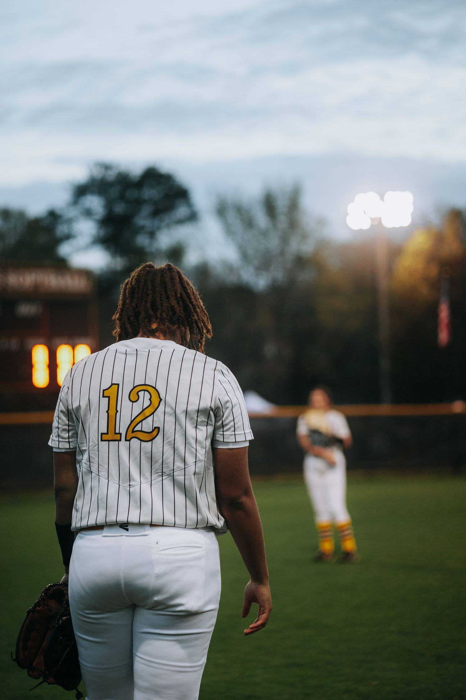 Player wearing jersey number 12 stands on softball field at dusk.