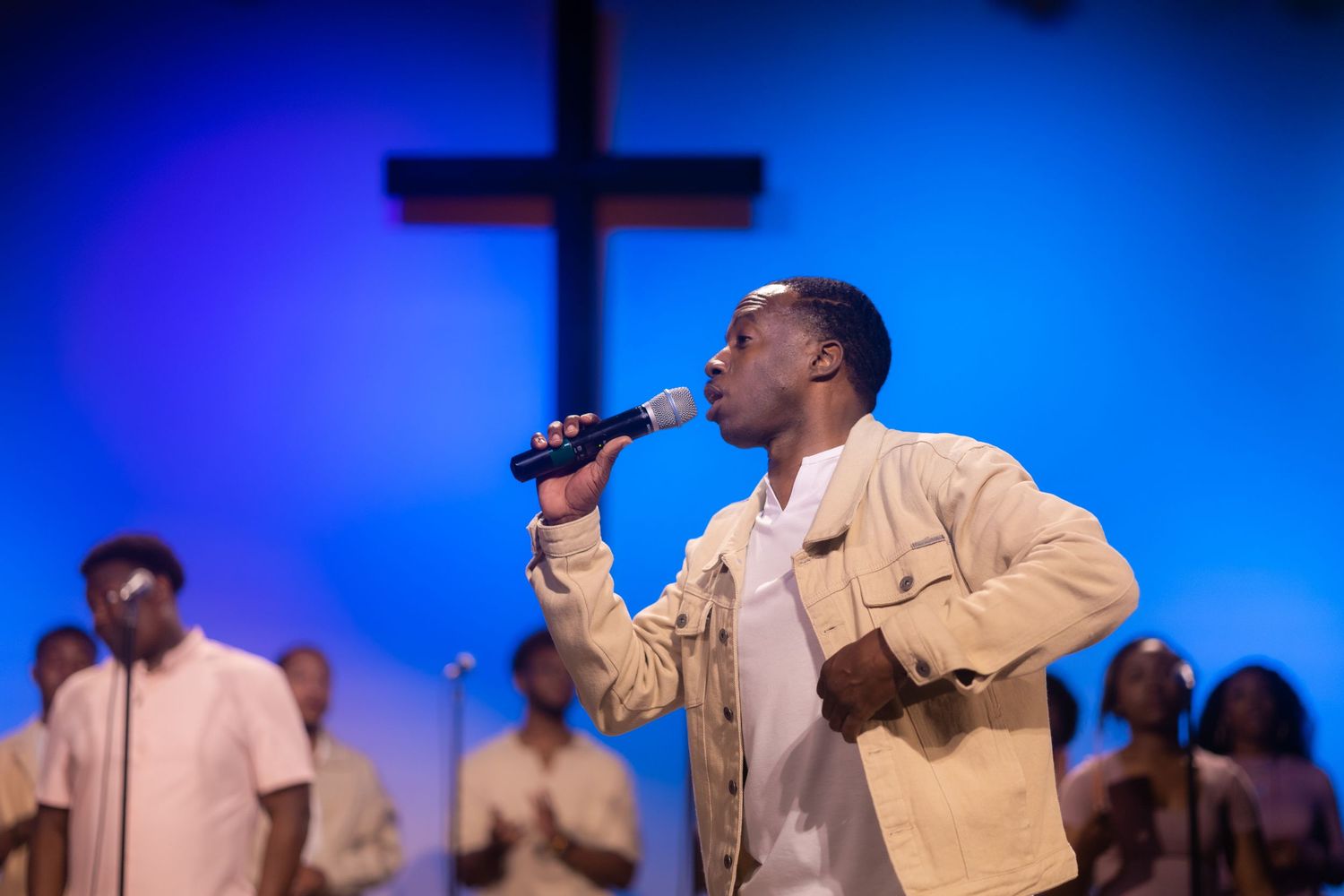 A performer in a beige jacket sings with emotion in front of a blue-lit cross.