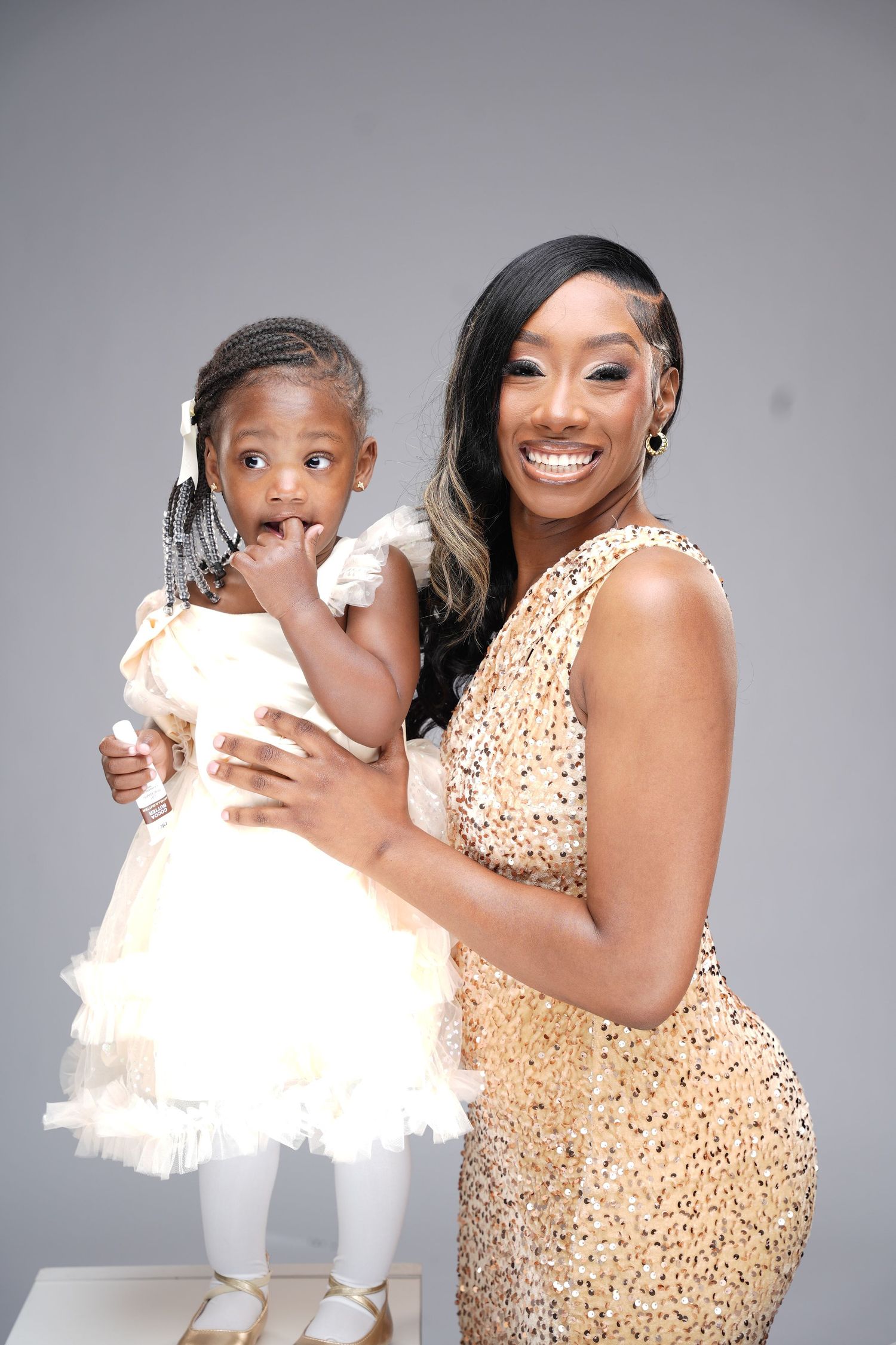 Mother and daughter in matching sparkly dresses share joyful moments during a professional photo shoot against grey backdrop.