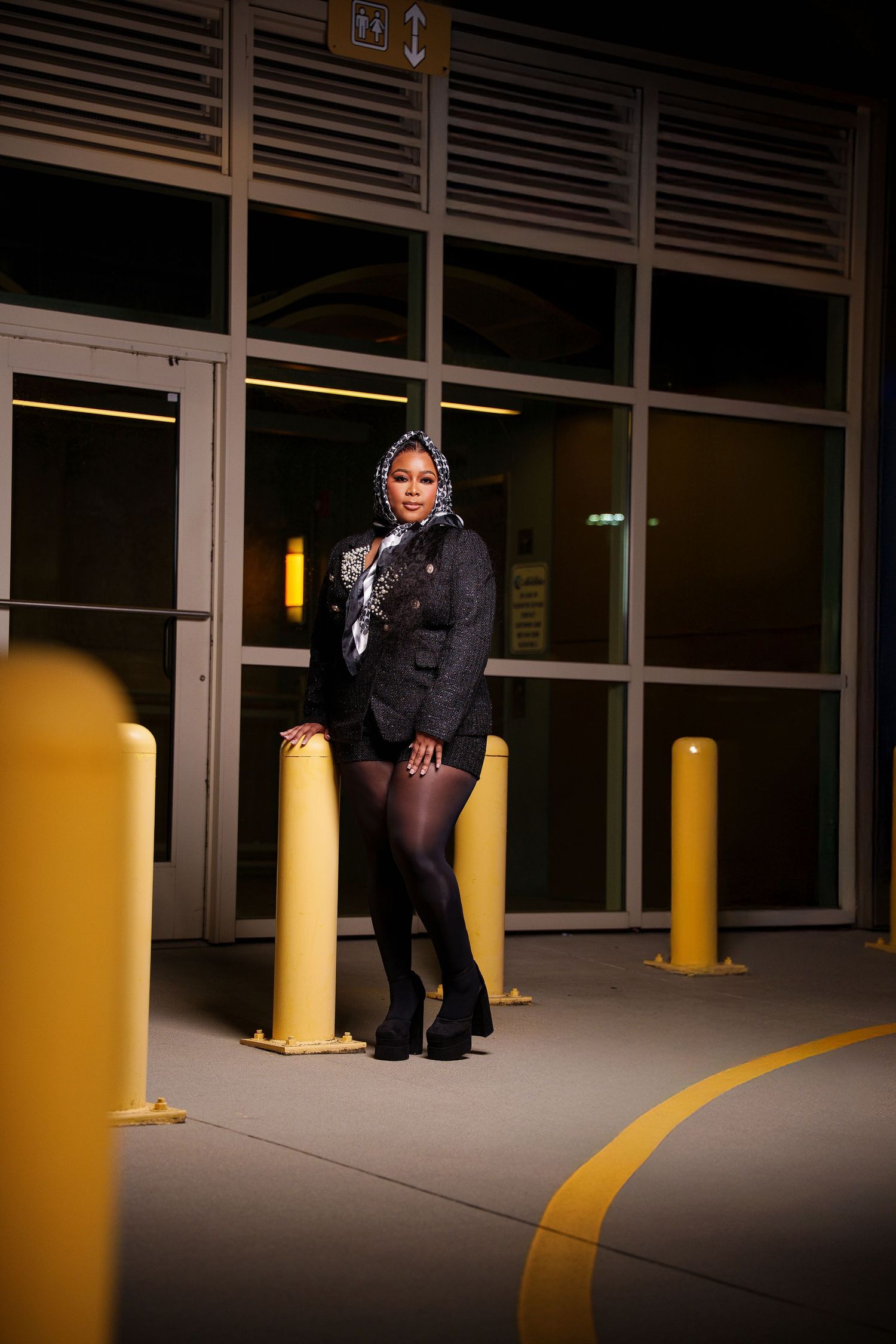A person in dark clothing crouches in a dimly lit parking garage with yellow poles and lines on the floor.