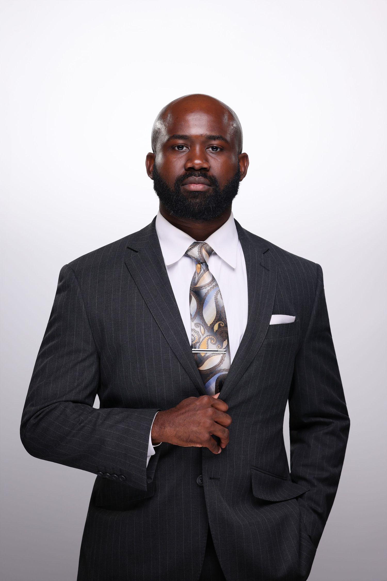 A confident bald Black businessman with a beard wearing a dark pinstripe suit and patterned tie poses professionally.