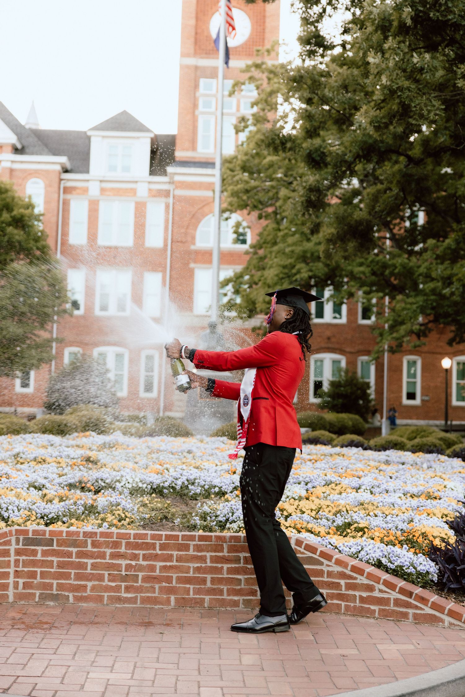 Graduate in red jacket celebrates with champagne in front of brick university building and colorful flower garden.