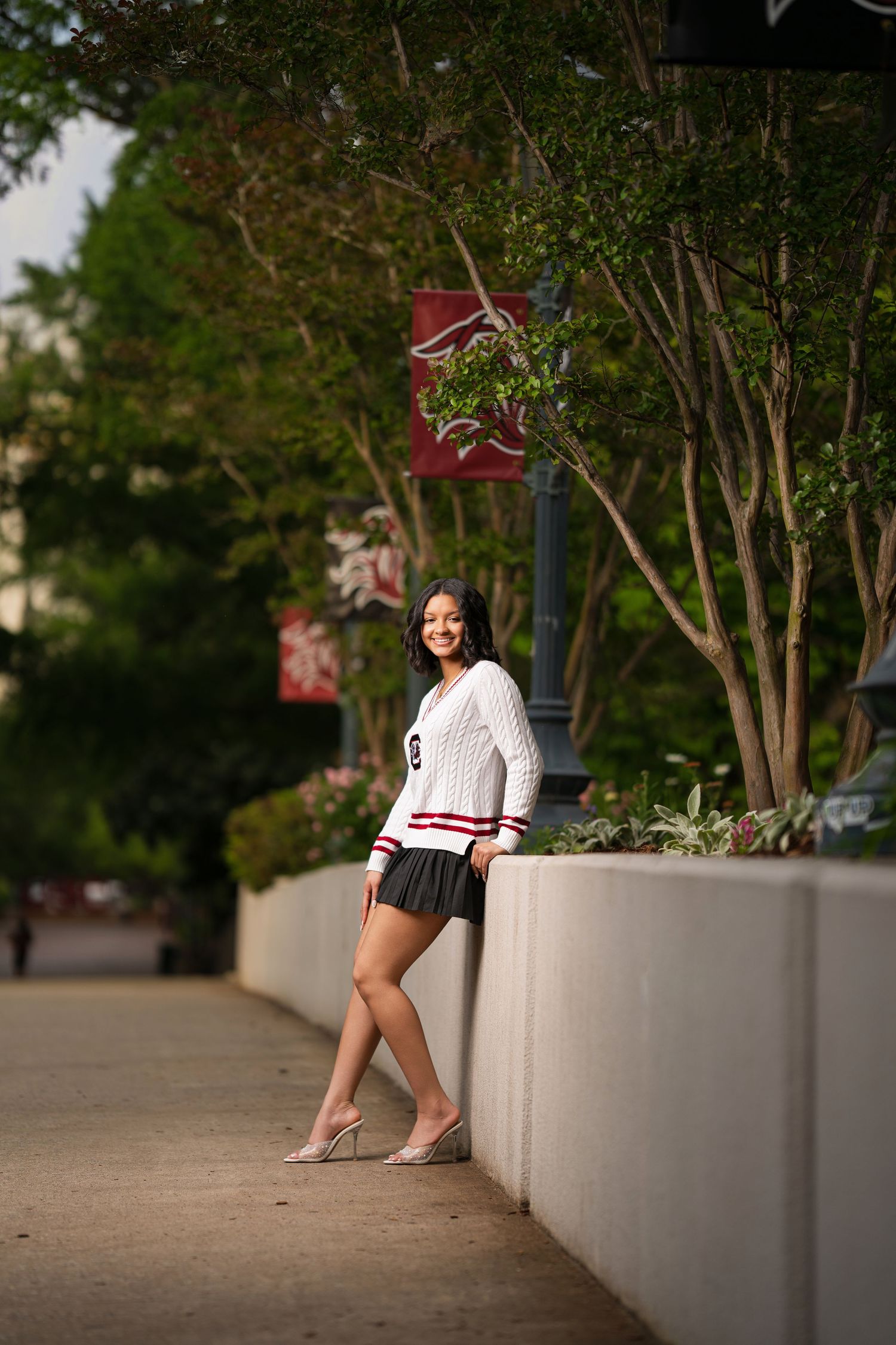 Young woman in white varsity sweater and mini skirt poses confidently along a tree-lined campus walkway.