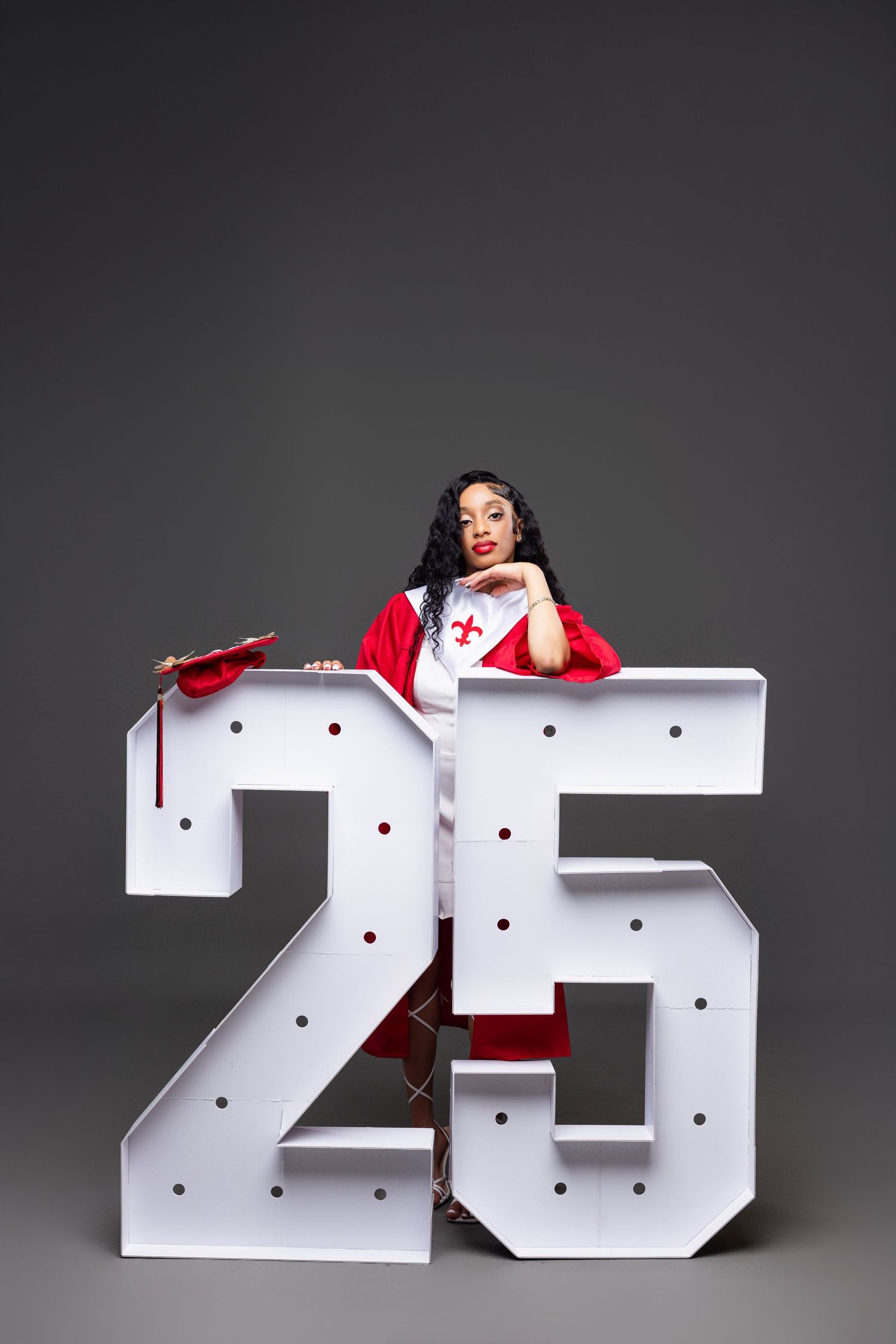 Graduate posing with large decorative number 25 sign in white and red against dark background.