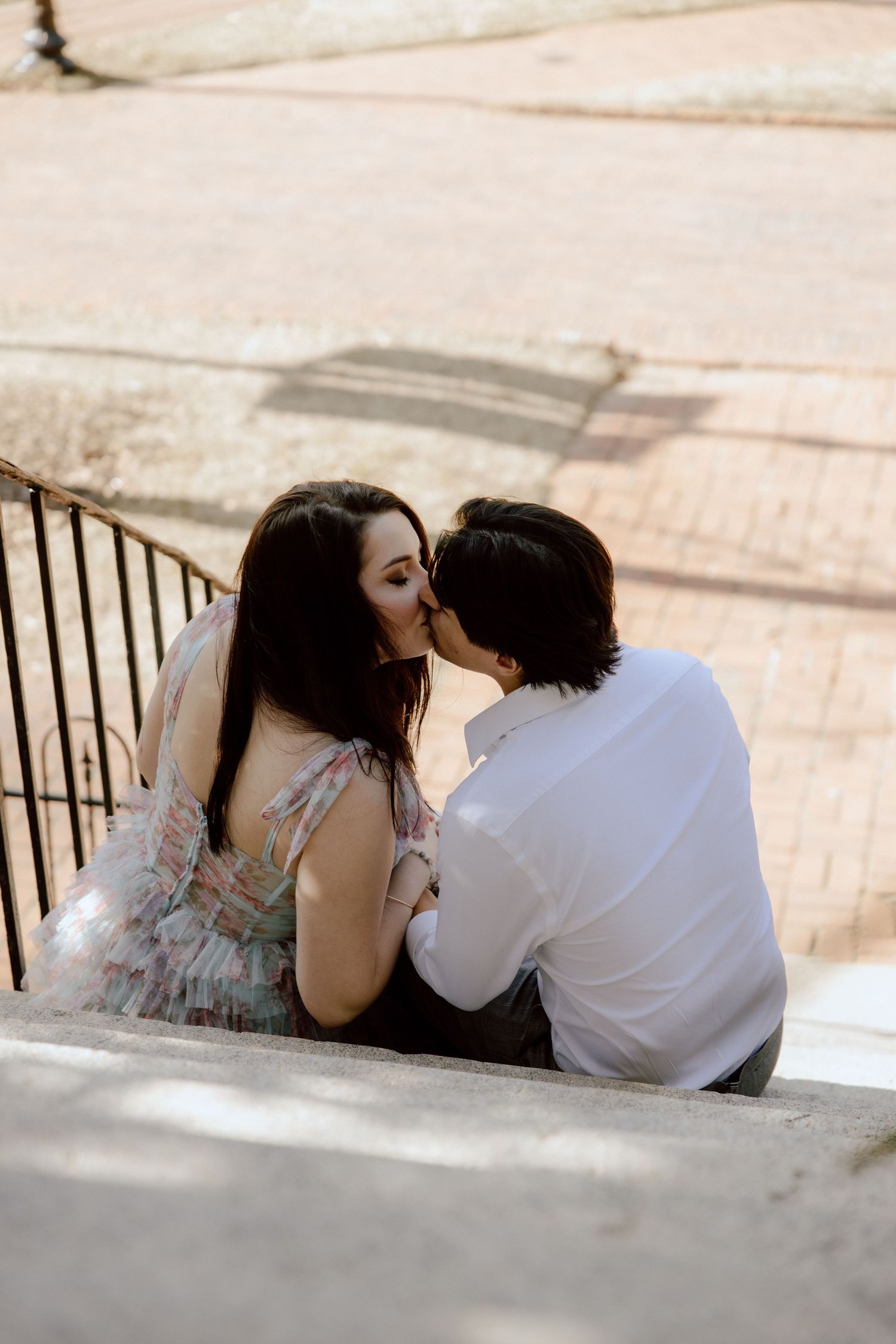 A romantic couple shares a kiss while sitting on concrete steps outdoors.