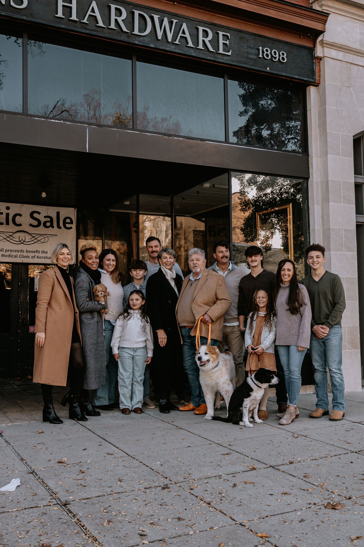 Large family group poses with two dogs outside historic hardware store.