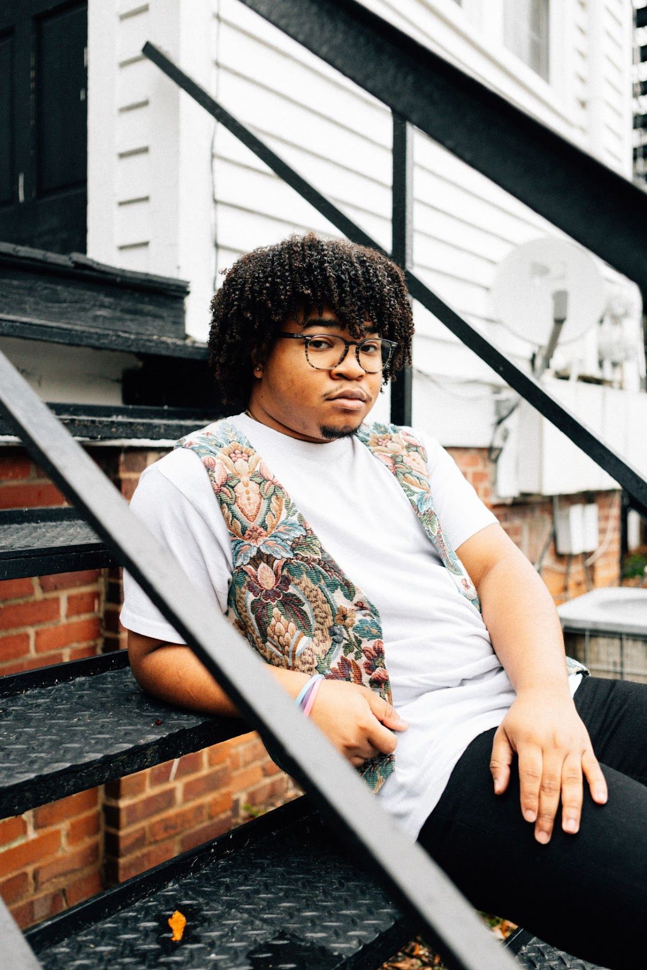 A person in glasses and white t-shirt with tattoos sits on outdoor steps against a brick wall.