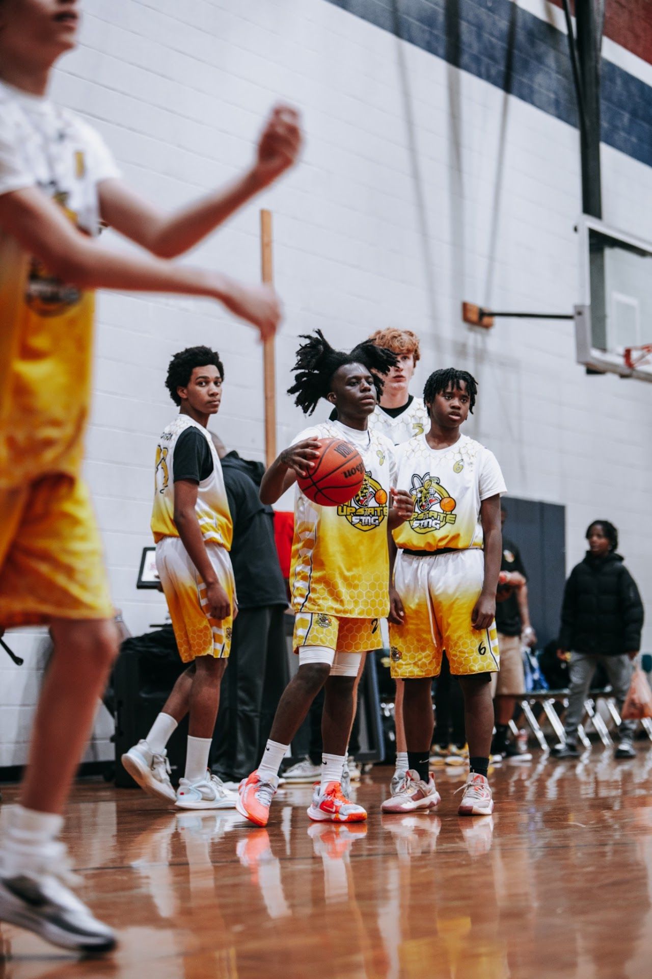 Basketball players in yellow and white uniforms sit along a gymnasium wall during a break.