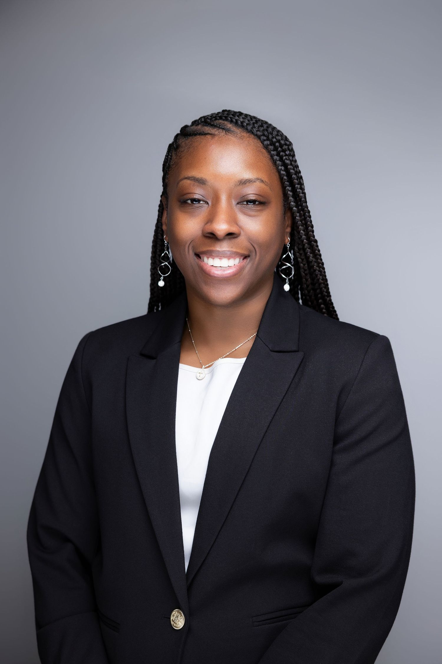 Professional headshot of a smiling woman in a black blazer with braided hair against a gray background.