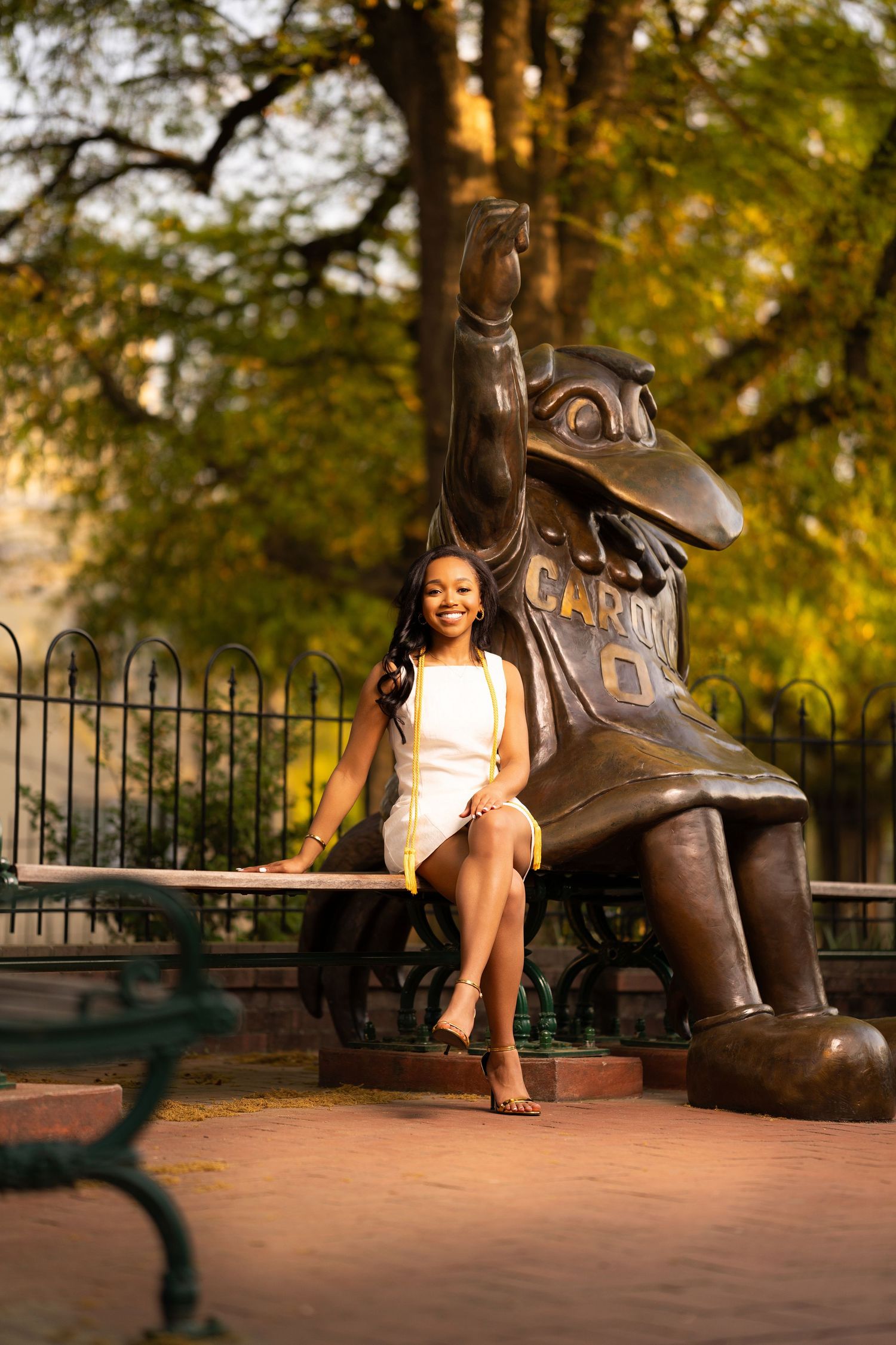 A smiling young woman in a white dress sits beside a Gamecock bronze mascot statue outdoors at the University of South Carolina.