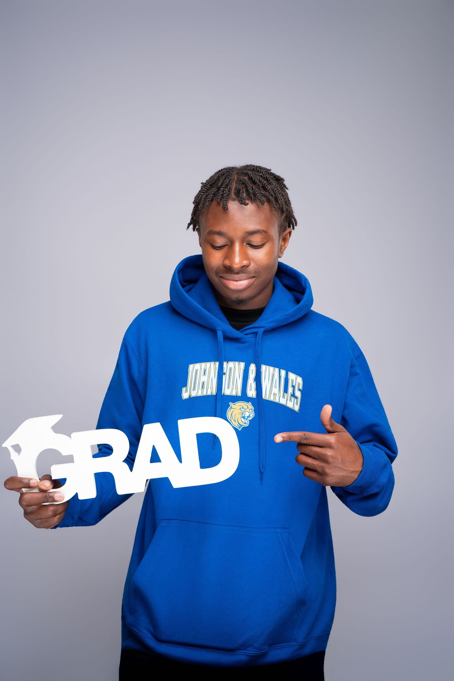 Young man in blue Johnson & Wales hoodie smiling and pointing at a white GRAD sign for graduation photos.