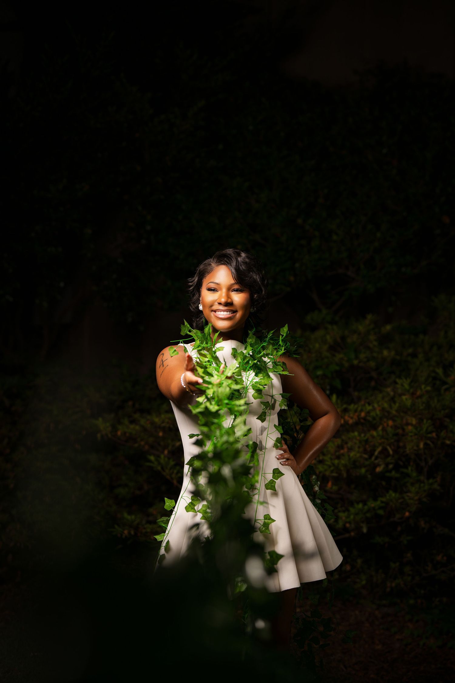 Young Black woman in white dress smiling outdoors at night, holding green ivy leaves in a dramatic spotlight.