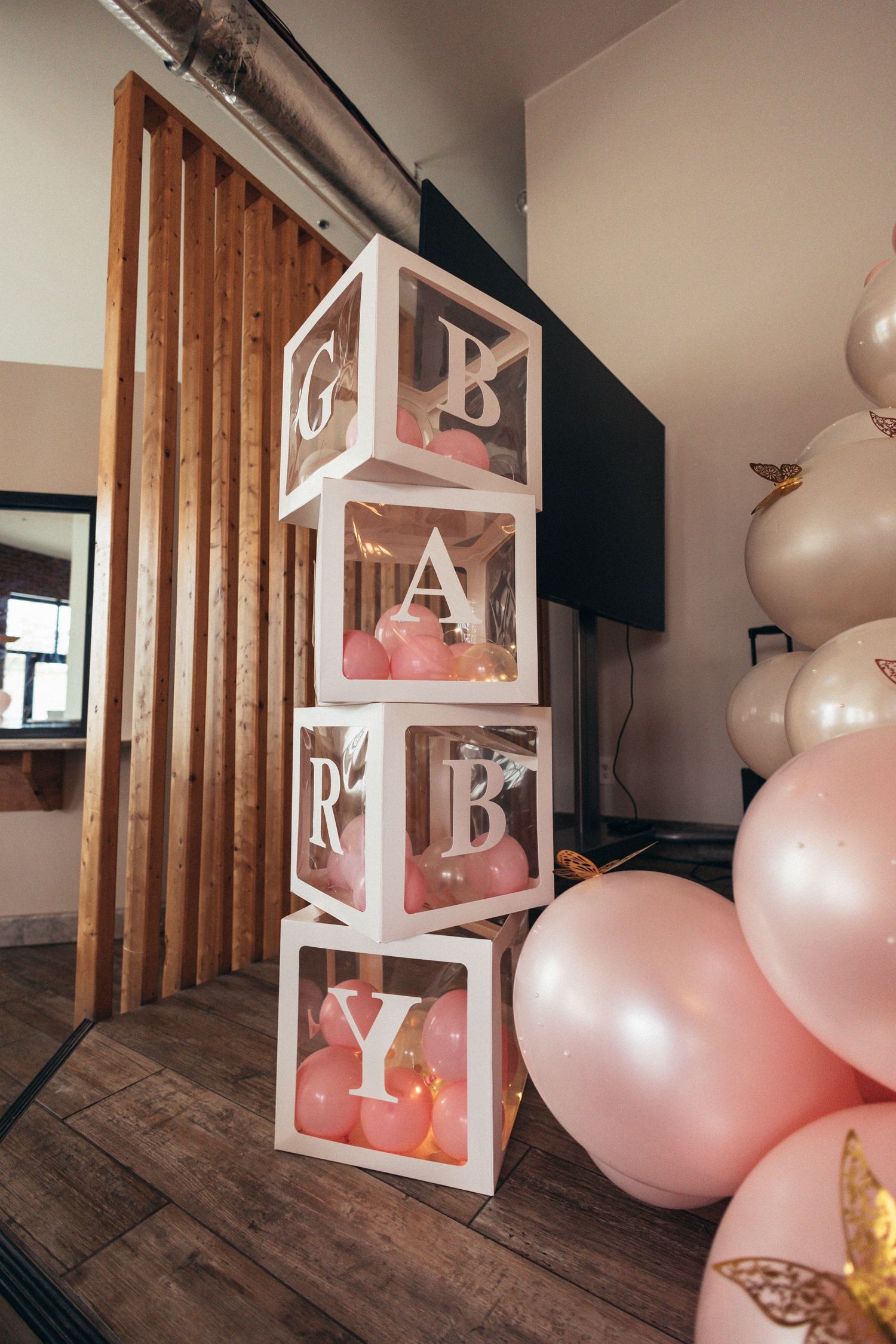 Pink baby shower decor with white letter blocks spelling BABY, filled with pink balloons, next to gold and pink balloon arrangement.