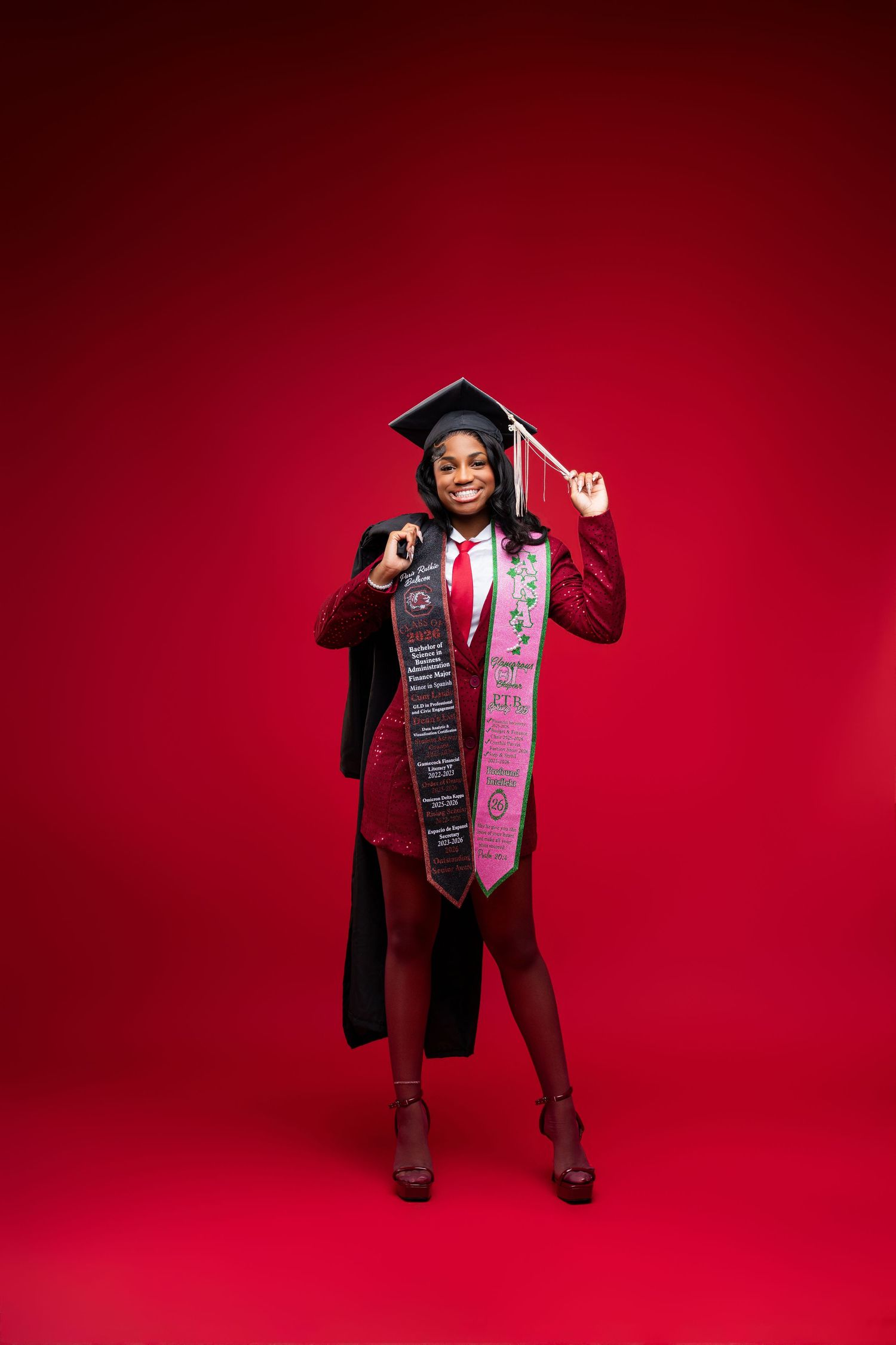 Smiling graduate in cap and gown with stoles poses confidently against a bold red background.