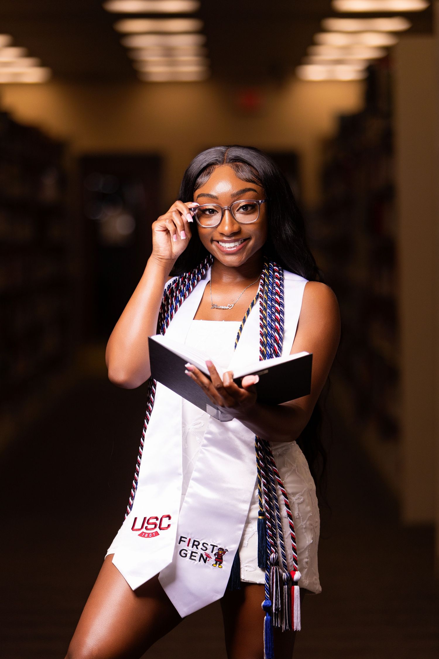 Student in graduation regalia adjusts glasses while holding book in library hallway.