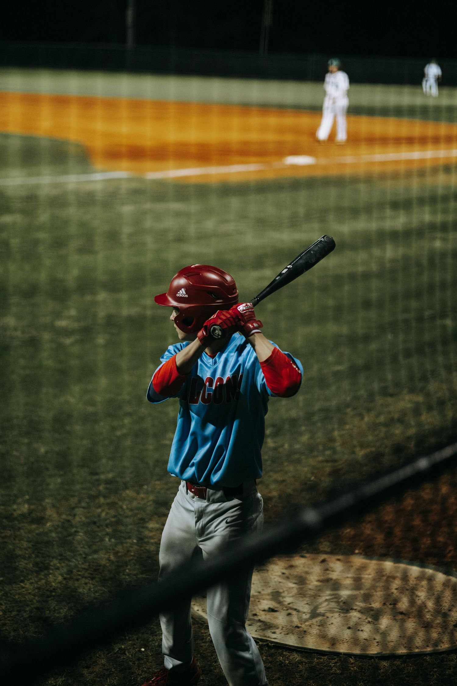 Baseball batter in red helmet and blue uniform prepares to swing at night game.