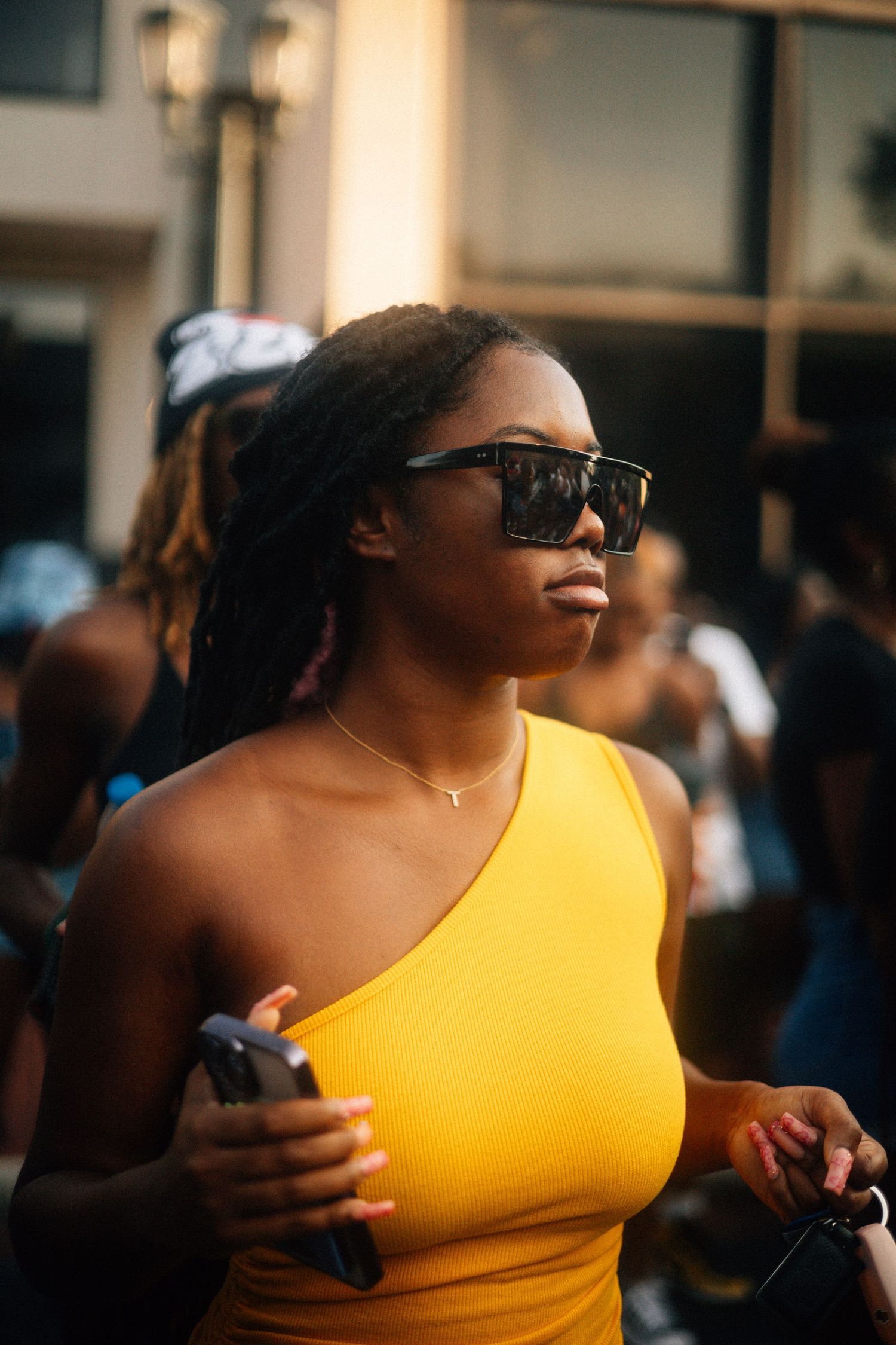 Person in a yellow one-shoulder top and sunglasses standing outdoors on a sunny day while holding a phone.  USC Hip Hop Wednesday