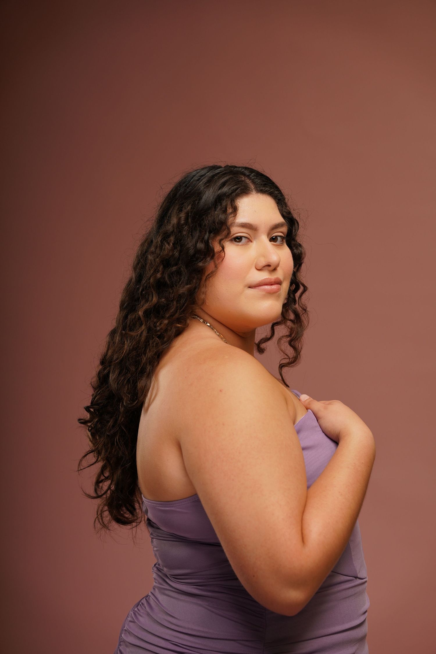 A person with long curly dark hair poses confidently in a purple sleeveless top against a brown background.