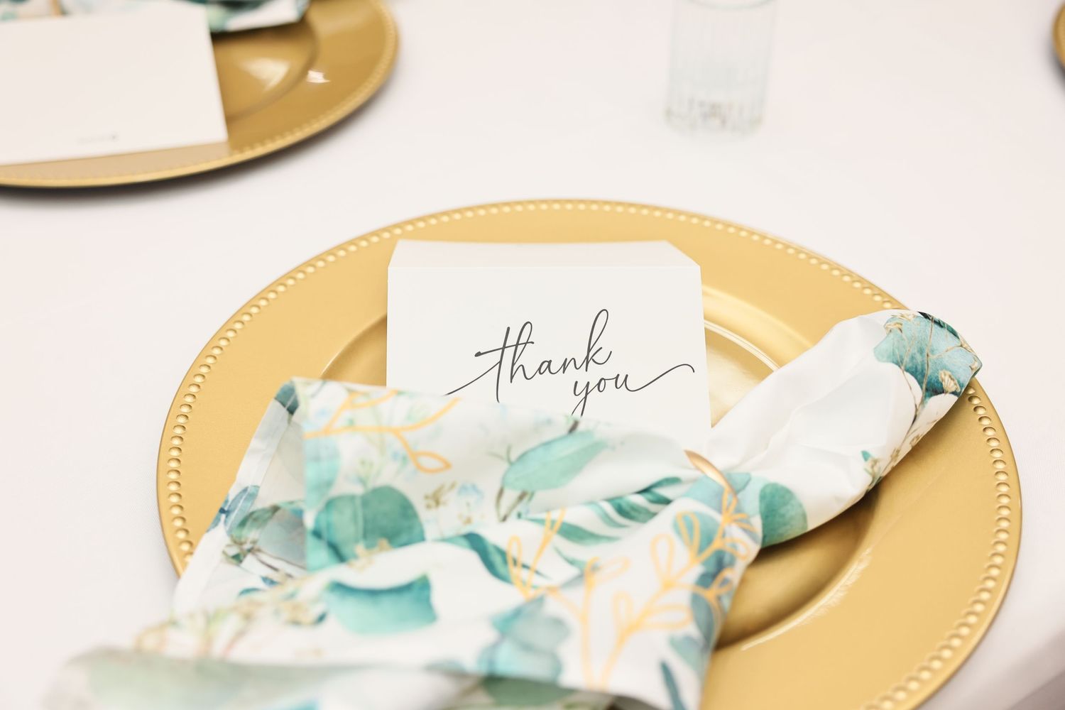 Gold charger plate with floral napkin and thank you card at elegant wedding table setting.