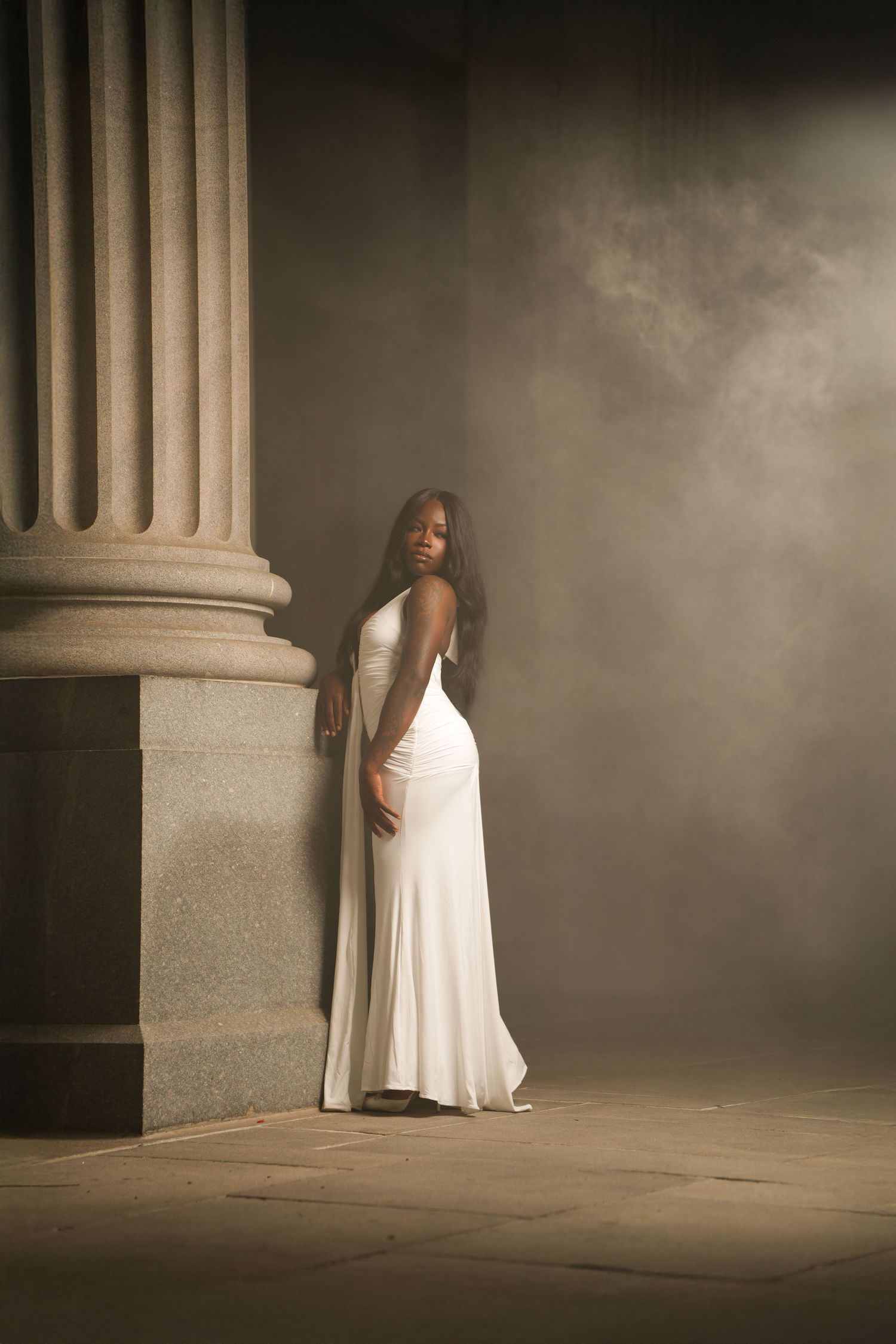 A Black woman in an elegant white gown poses beside a stone column in a dramatic, smoky atmosphere.