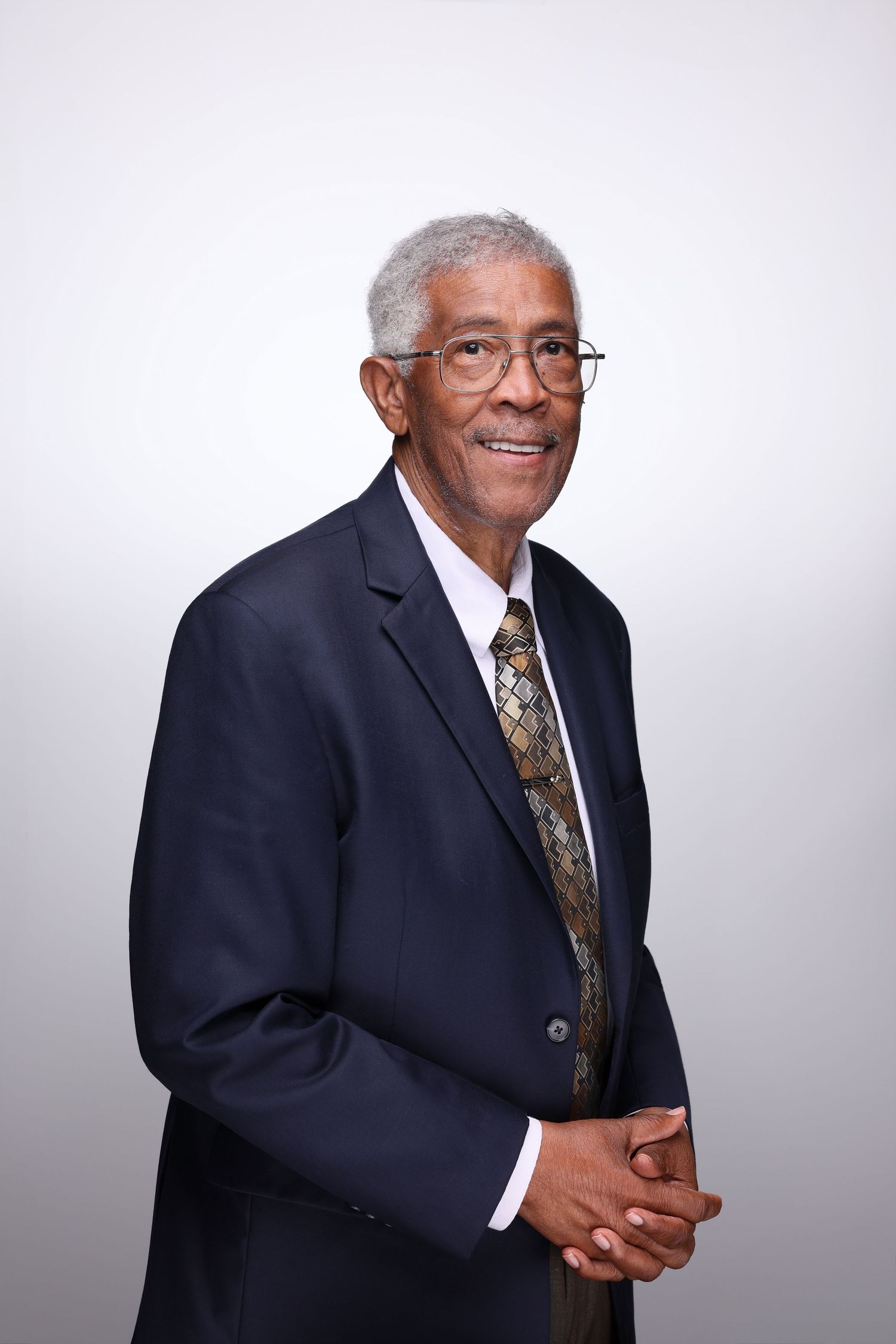 Elderly man in navy suit and gold tie smiling, wearing glasses, hands clasped, professional portrait on gray background.