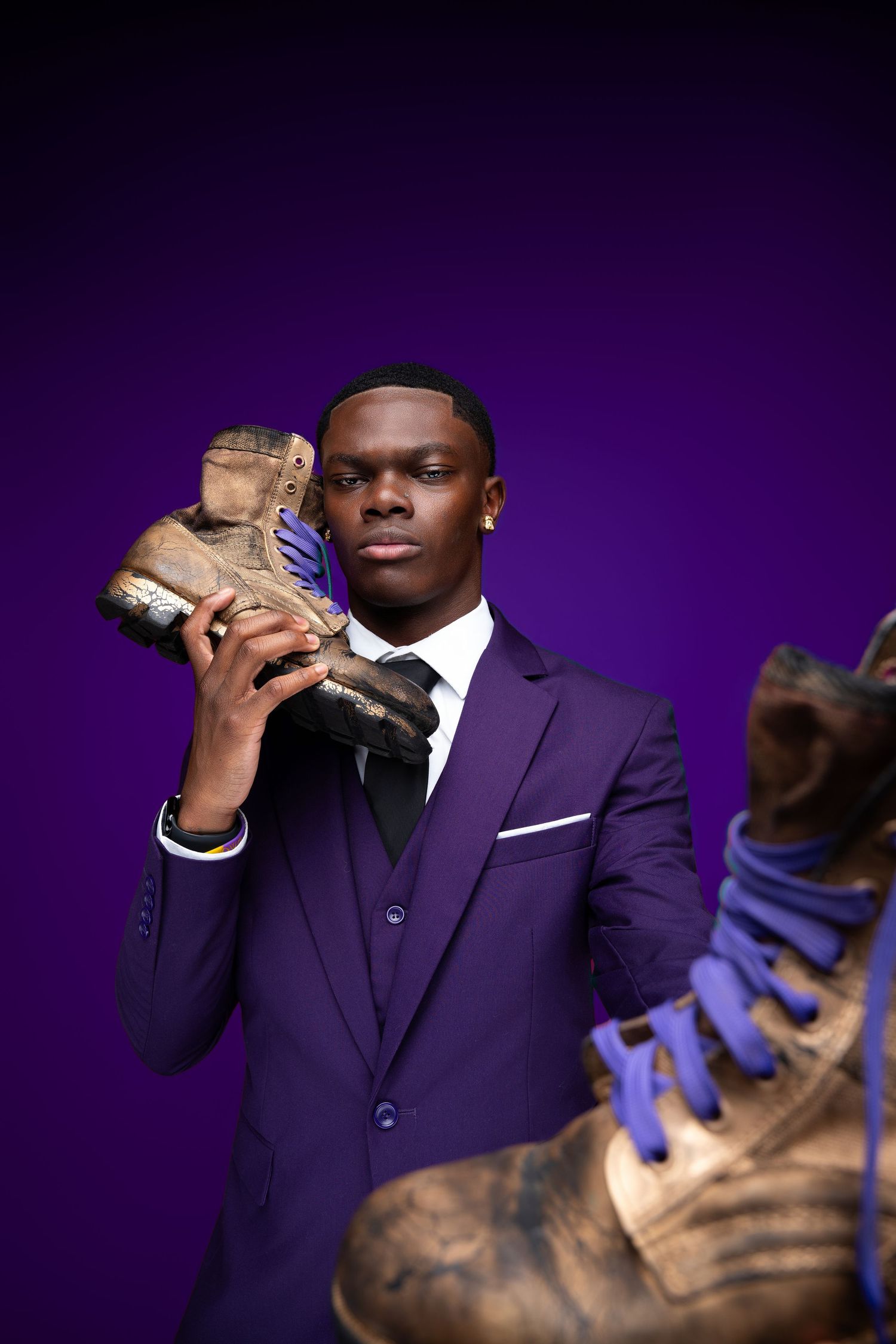 Young man in purple suit holding worn boots against purple background.