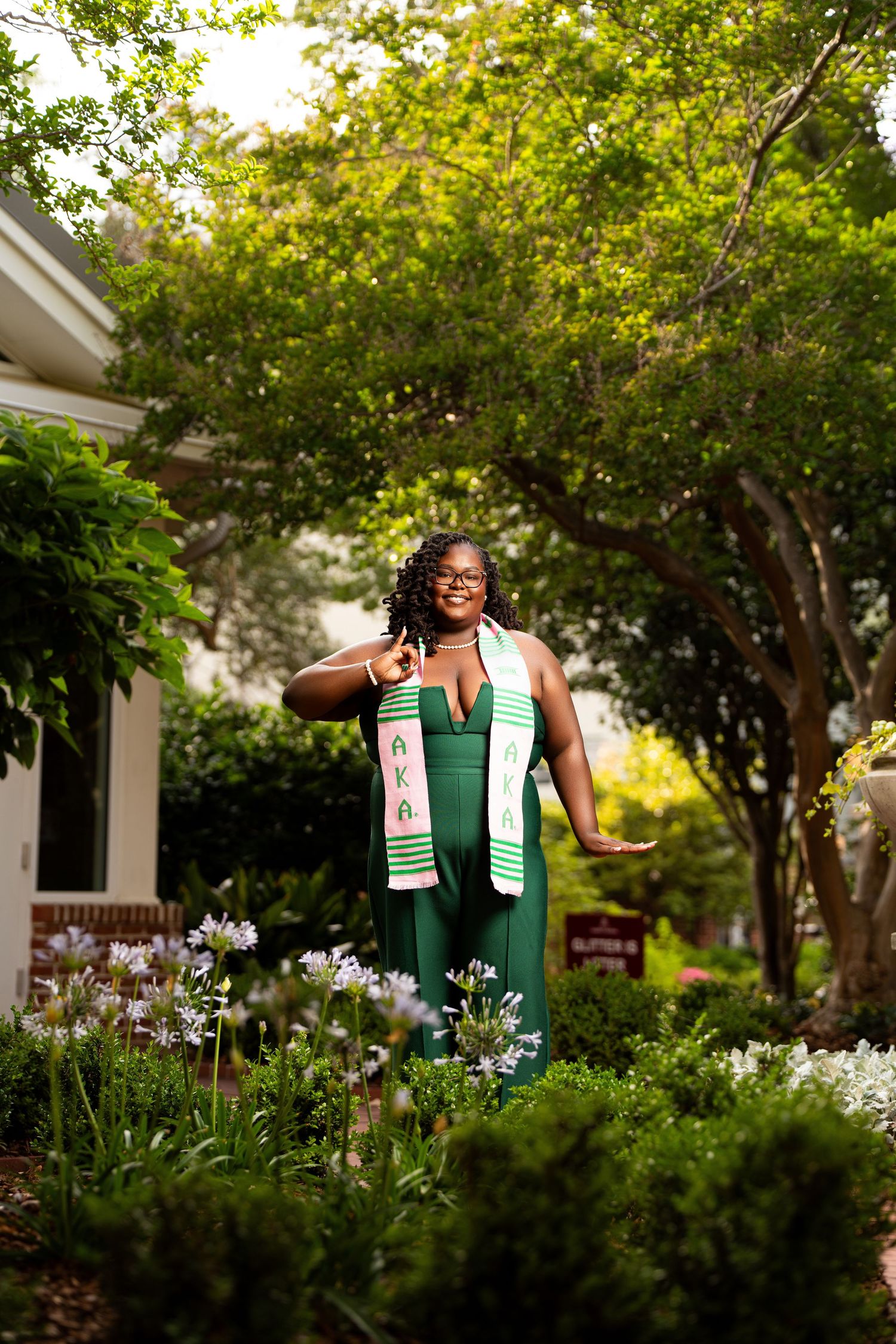 Woman in green gown with AKA graduation stole poses proudly in lush garden setting.