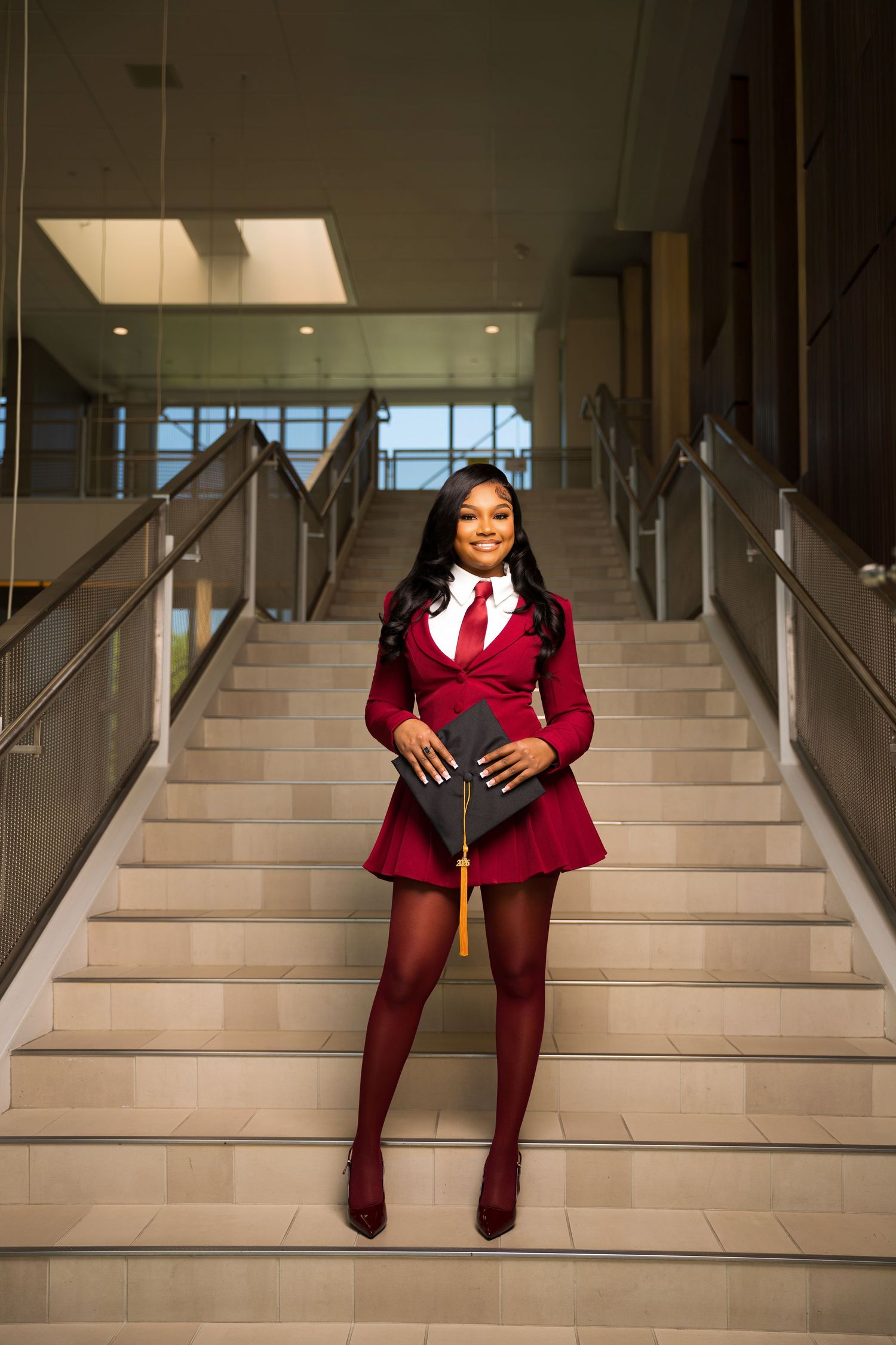 Young woman in red graduation attire holding cap on stairs, celebrating academic achievement.