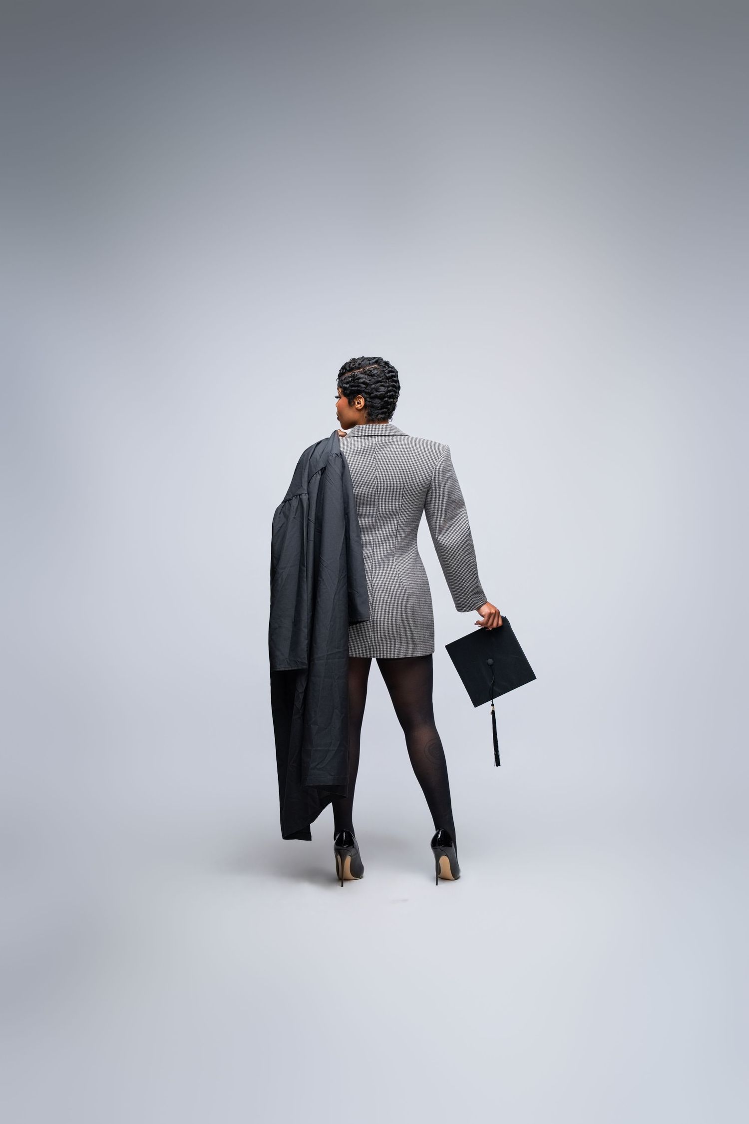 Graduate woman in blazer and heels holding cap and gown, rear view on gray background.