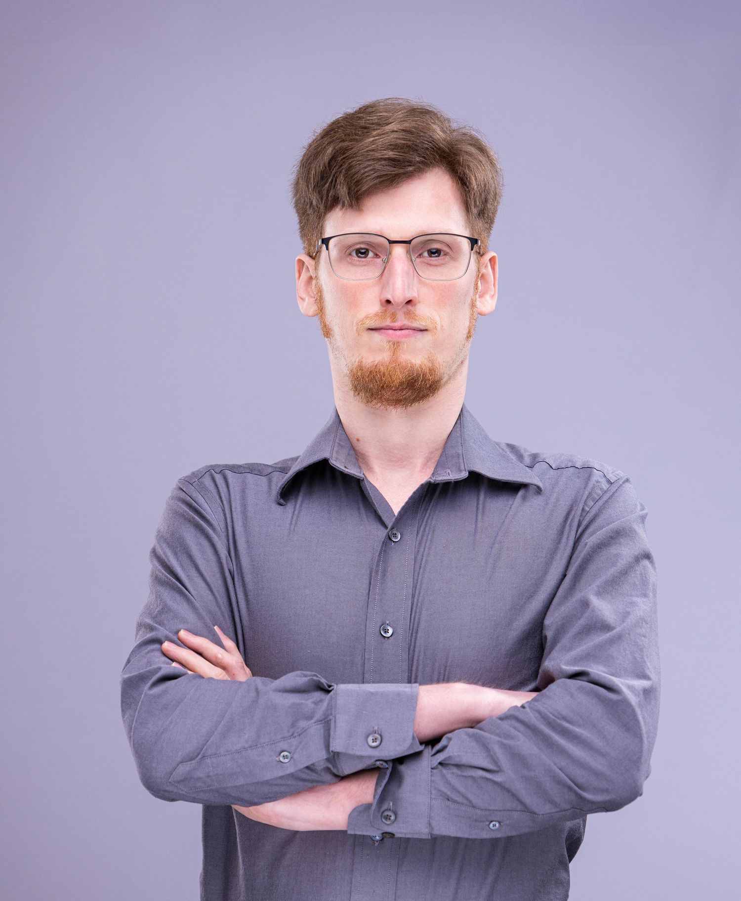 Professional headshot of person in gray button-down shirt with crossed arms against purple background.