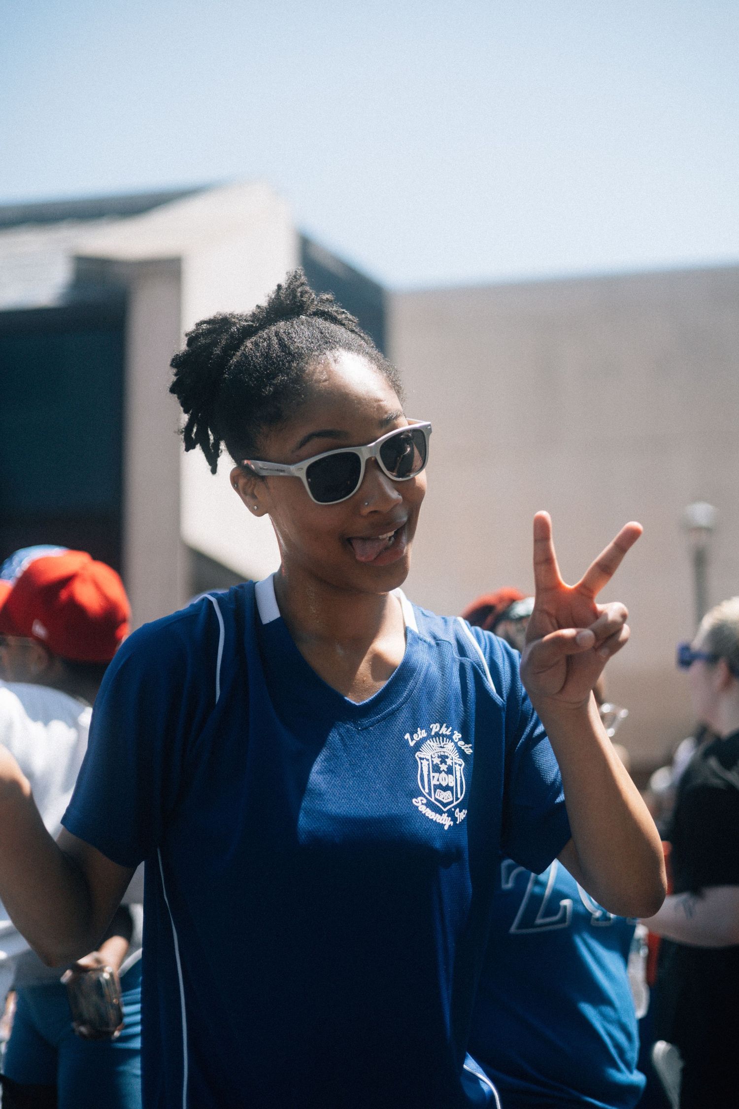 Someone in sunglasses wearing a blue t-shirt makes a peace sign gesture while smiling outdoors on a sunny day.  USC Hip Hop Wednesday