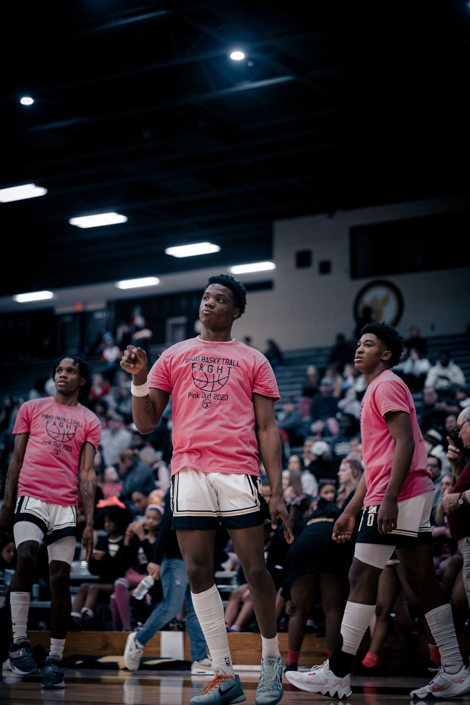 Basketball team in pink jerseys watching game from sideline in dark gymnasium.