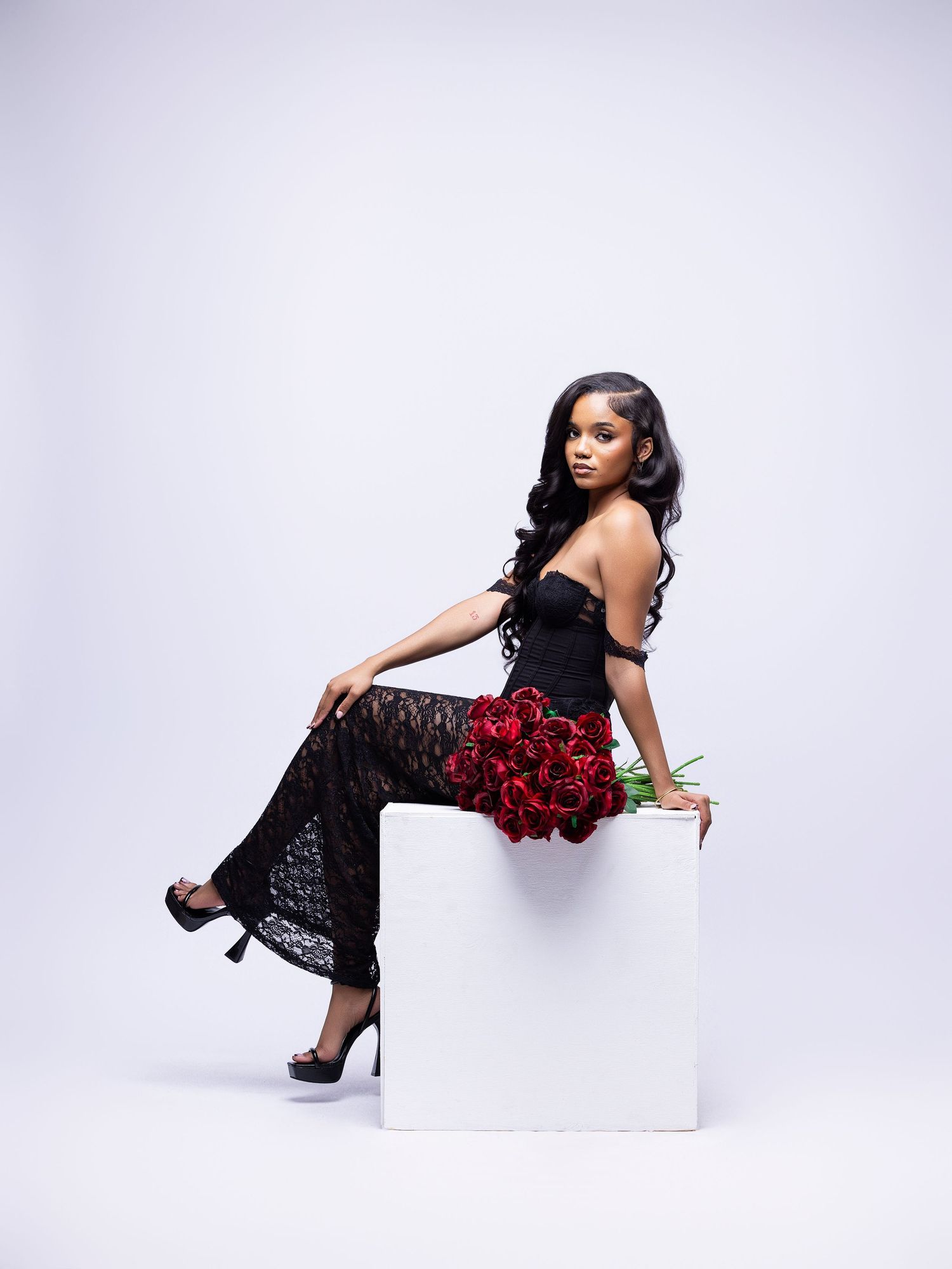 A glamorous figure in a black lace dress poses with red roses while seated on a white cube against a light background.