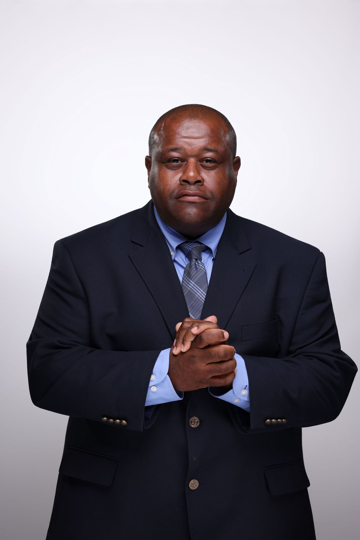 Professional Black businessman in dark suit and gray tie, hands clasped, serious expression, studio portrait.