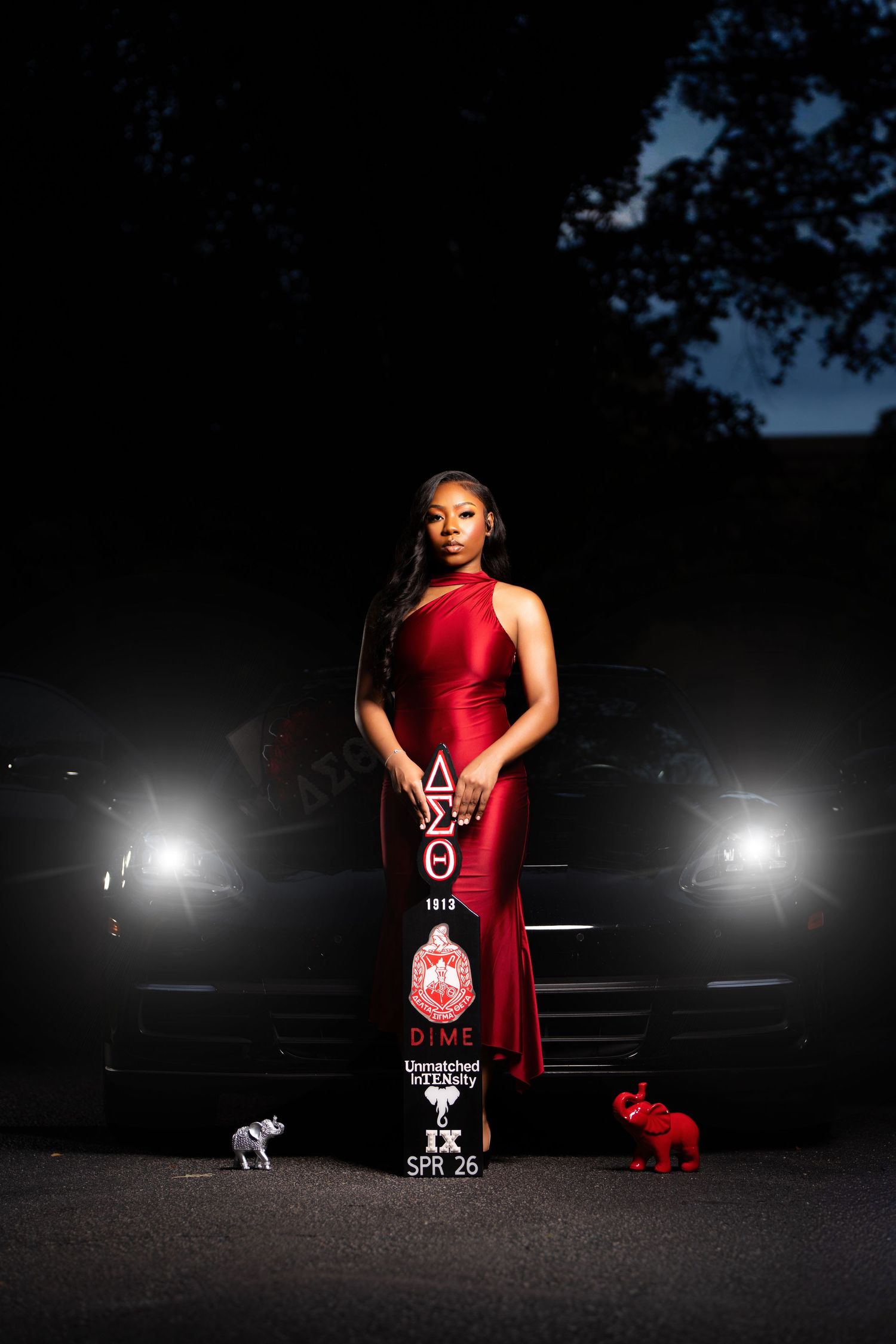 Woman in red gown poses before car headlights holding Delta Sigma Theta sorority paddle at night.