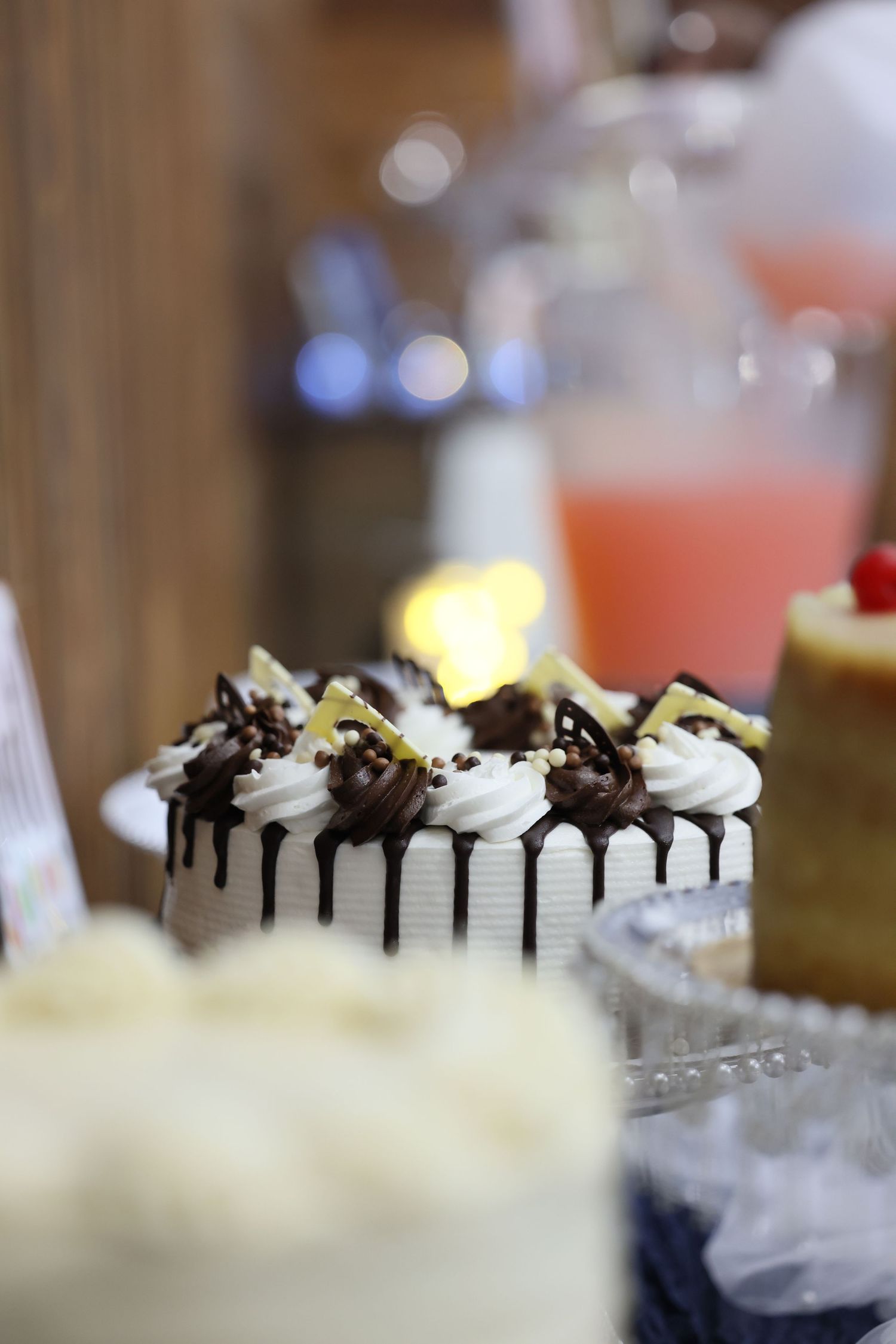 Elegant white cake with chocolate drizzle and cream rosettes on a dessert table with blurred background.