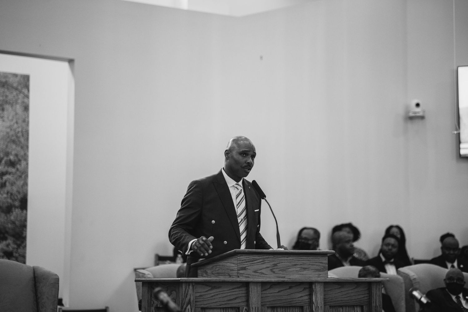 A black and white photograph shows a speaker addressing an audience in a meeting room setting.