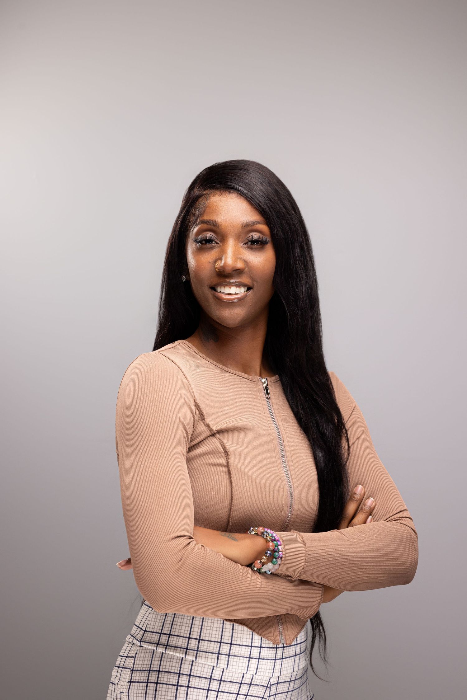 A confident young Black woman with long hair, wearing a beige top, stands with arms crossed and smiles at the camera.