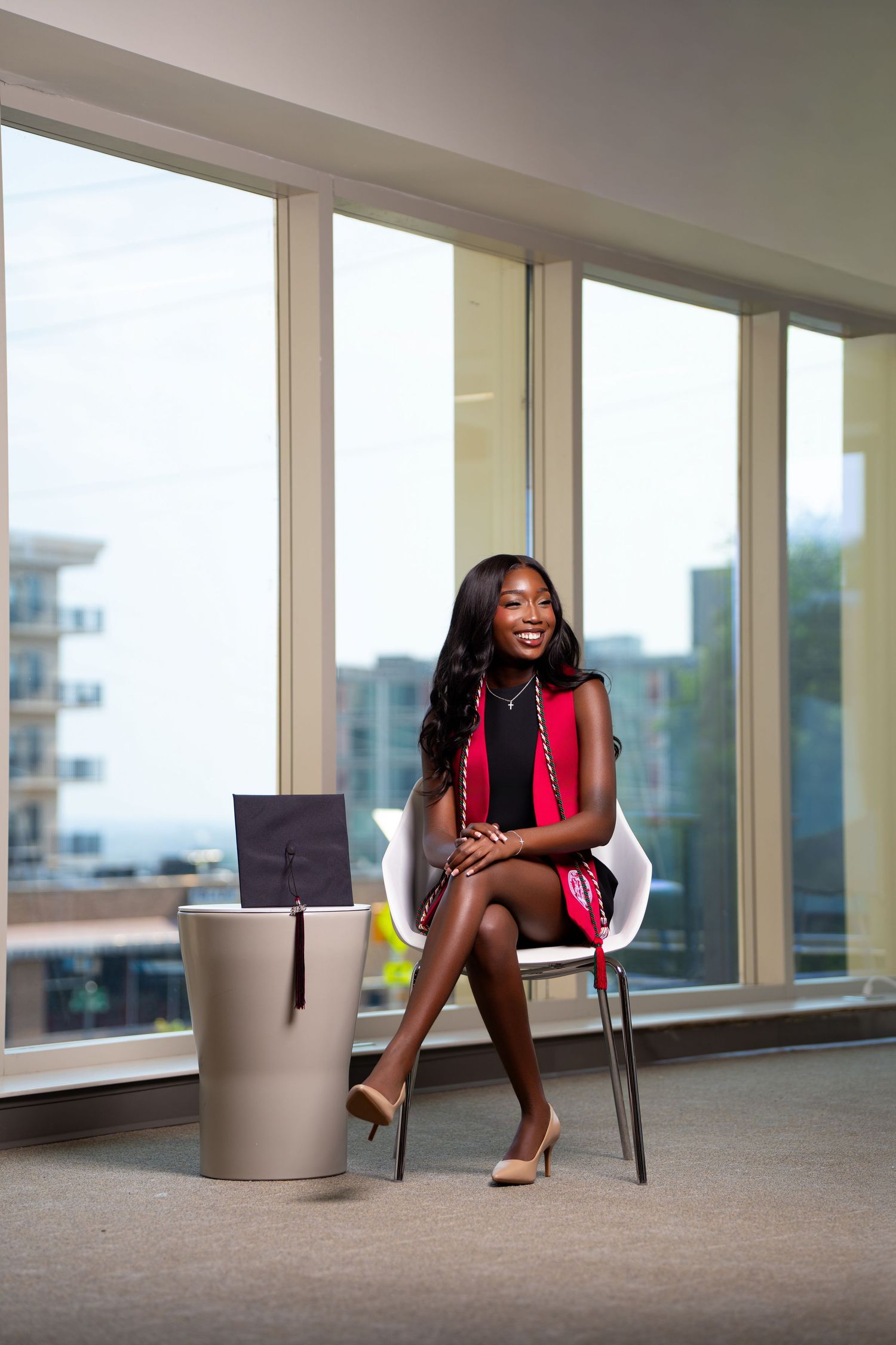 Young Black woman in red dress sits smiling in modern office with graduation cap beside her.