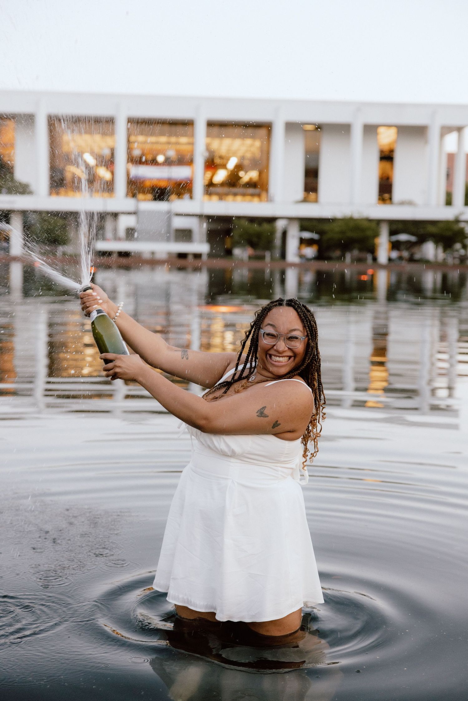 Woman in white dress celebrating in water, spraying champagne bottle, smiling joyfully outdoors.